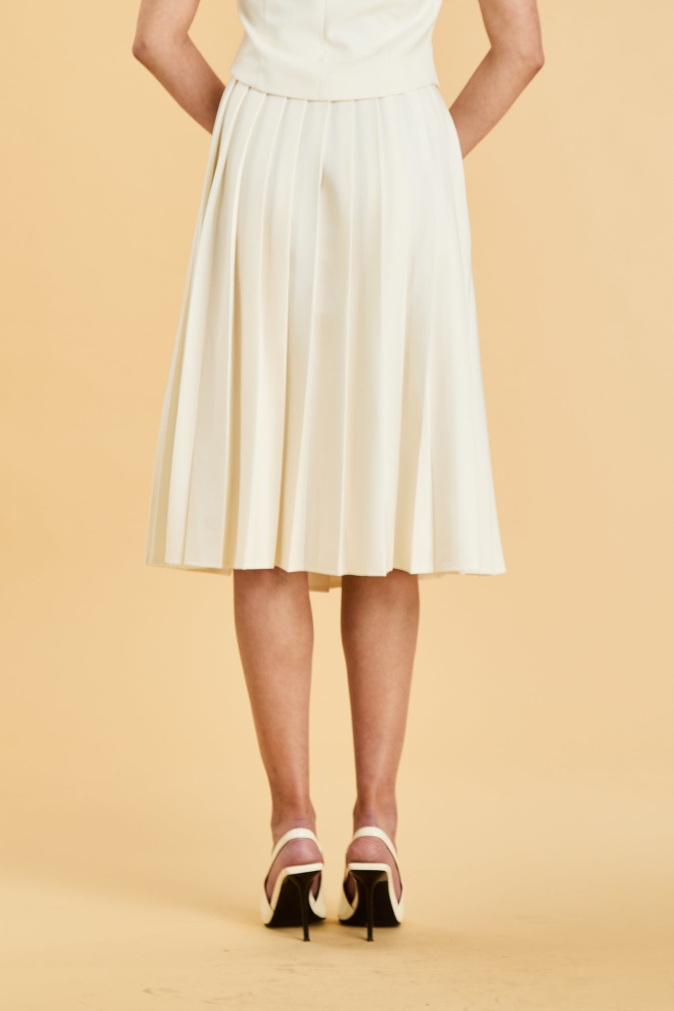 Close up of a woman wearing a White colored Triplicity Alice top and White Triplicity pleated midi skirt against a beige background back view