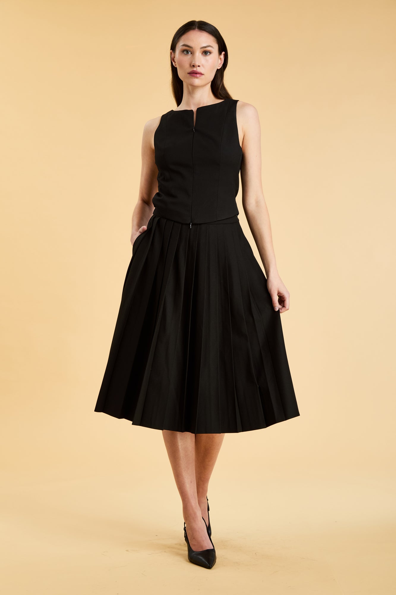 Woman wearing a black sleeveless top on a pleated Black midi skirt on a beige background