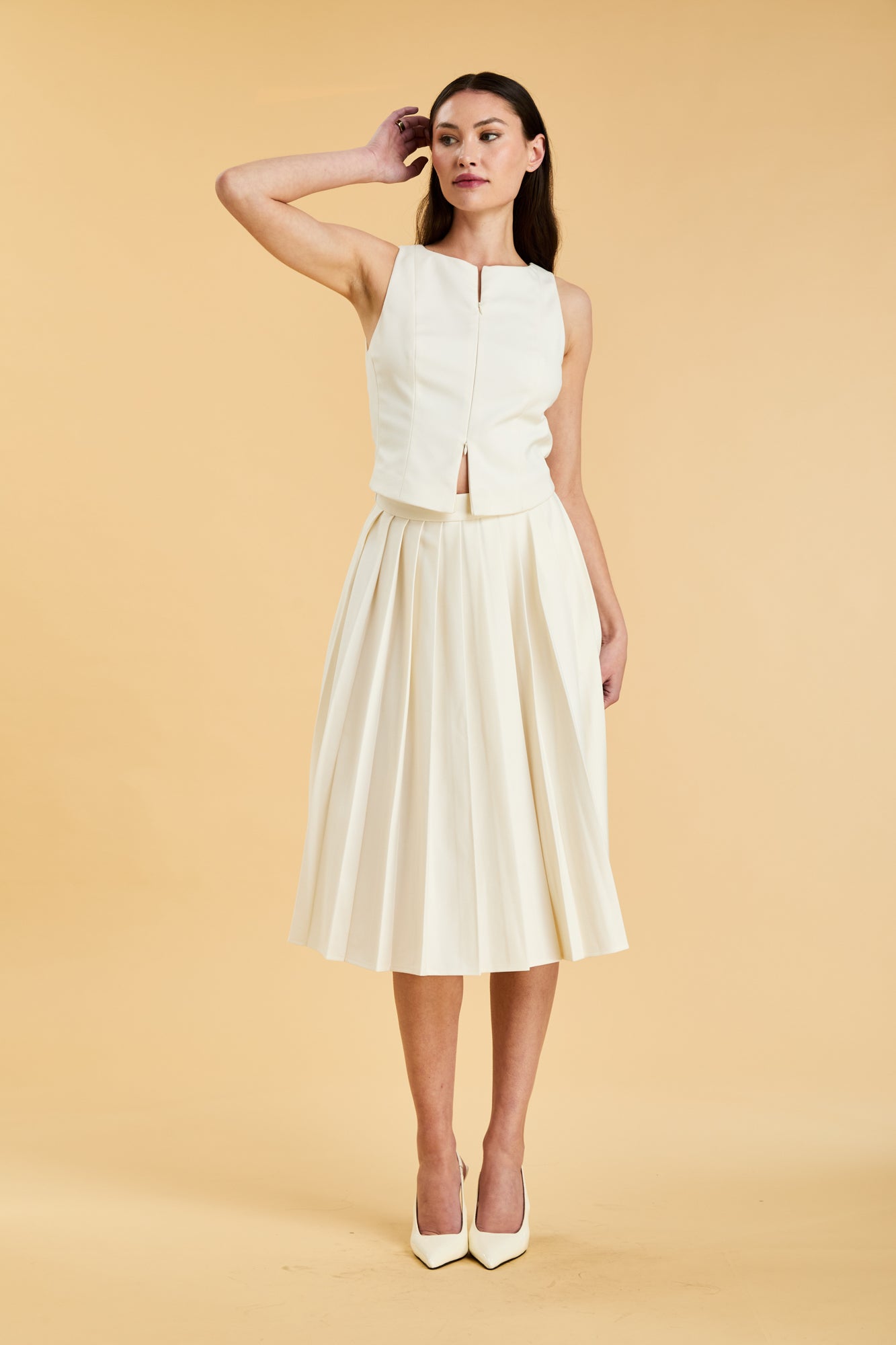 Woman wearing a White colored Triplicity double ended zipper Alice top and White Triplicity pleated midi skirt against a beige background front view full body