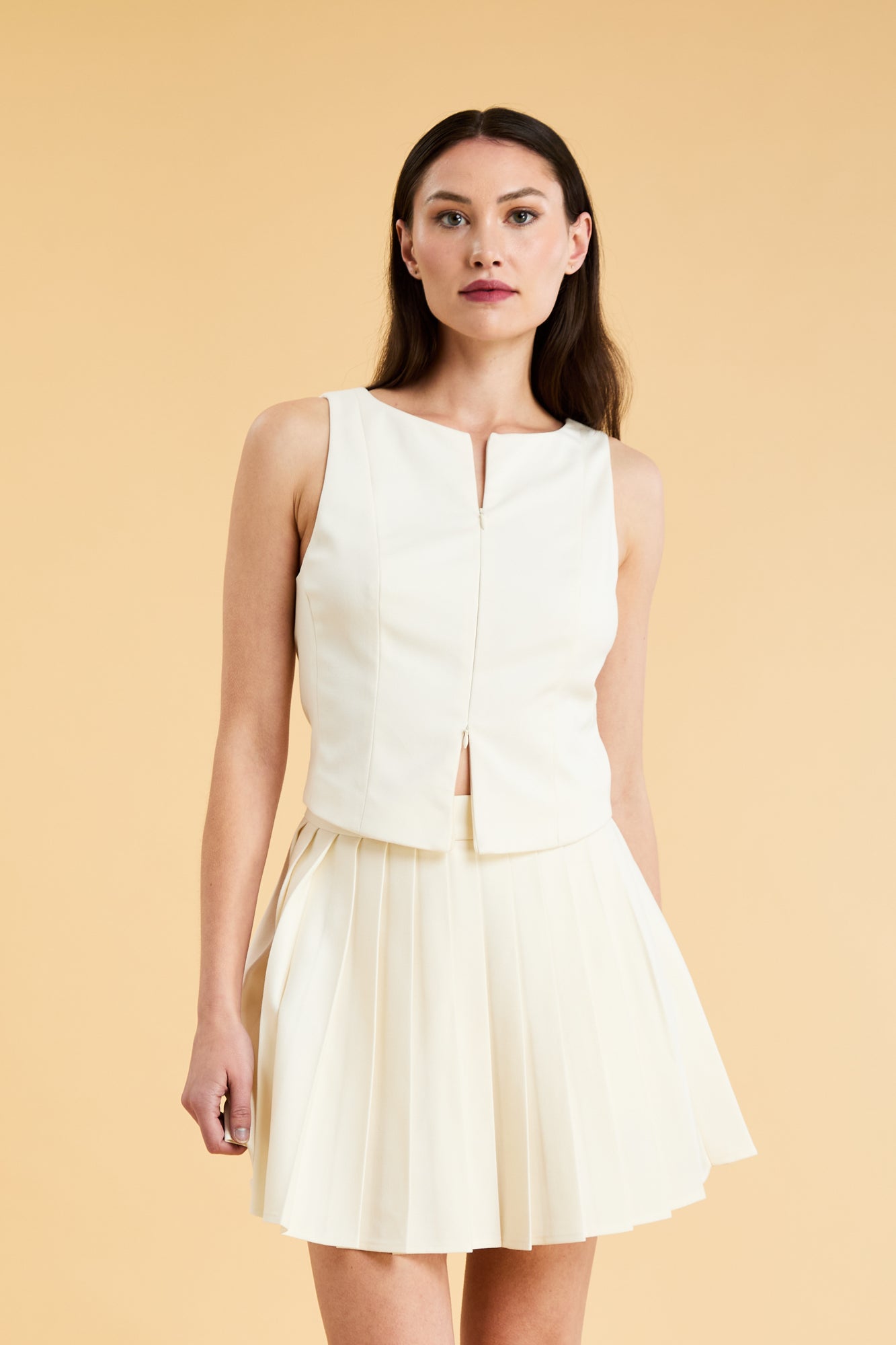 Woman wearing a White Triplicity double ended zipper sleeveless top and Triplicity White pleated skirt against a beige background