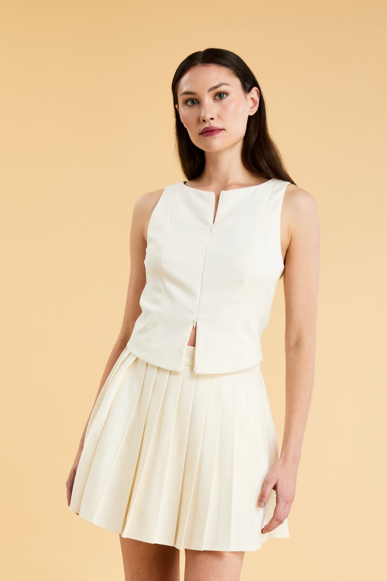 Woman wearing a White Triplicity double ended zipper sleeveless top and Triplicity White pleated skirt against a beige background