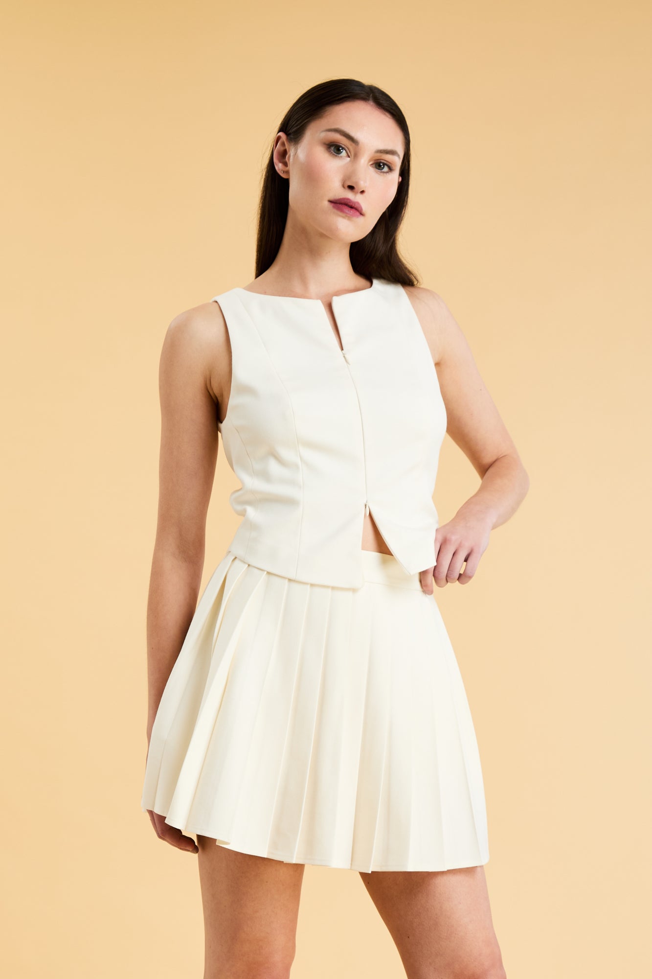 Woman wearing a White Triplicity double ended zipper sleeveless top and Triplicity White pleated skirt against a beige background
