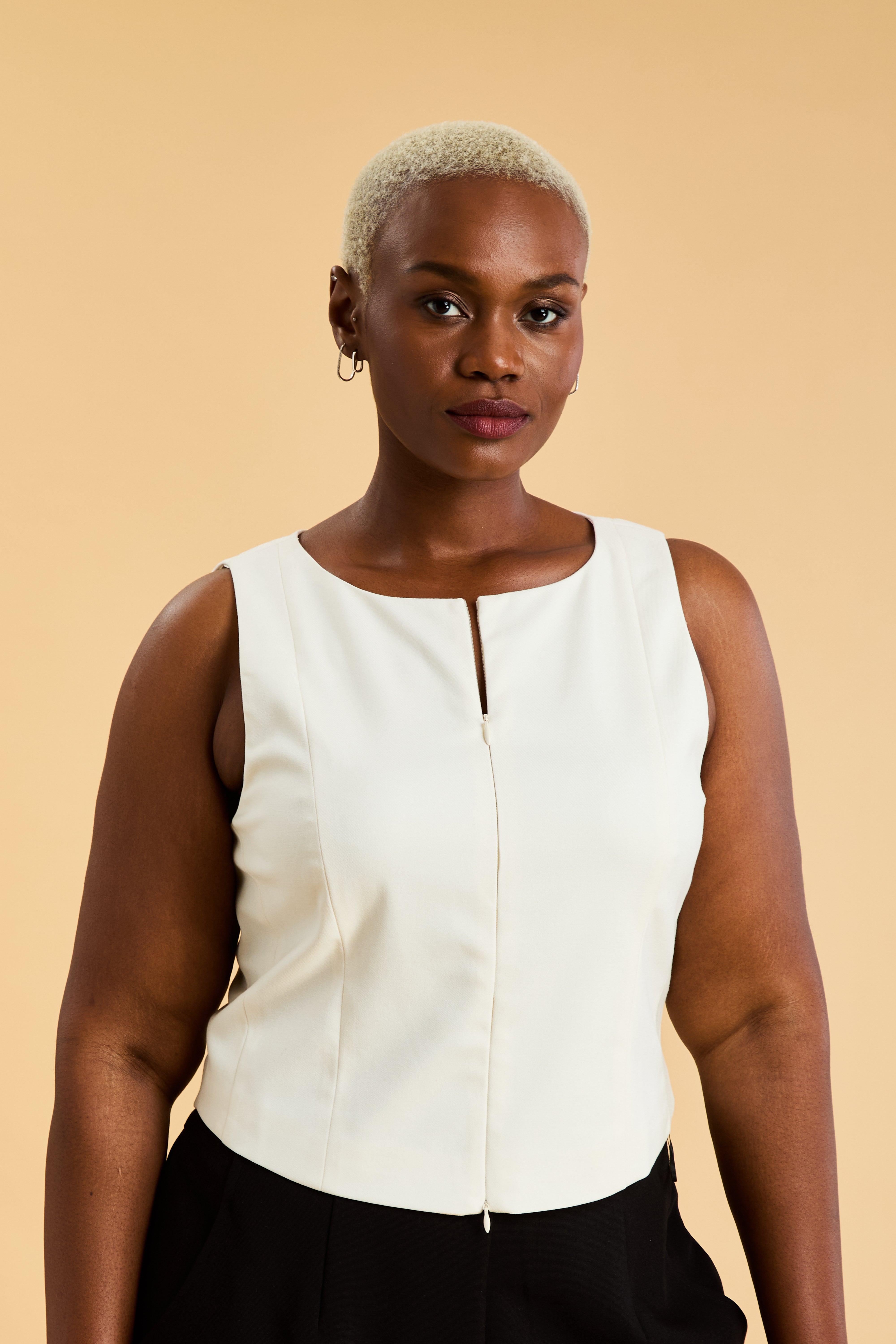 Woman wearing a White Triplicity sleeveless Alice double ended zipper top and Almond colored pants against a beige background close up front view