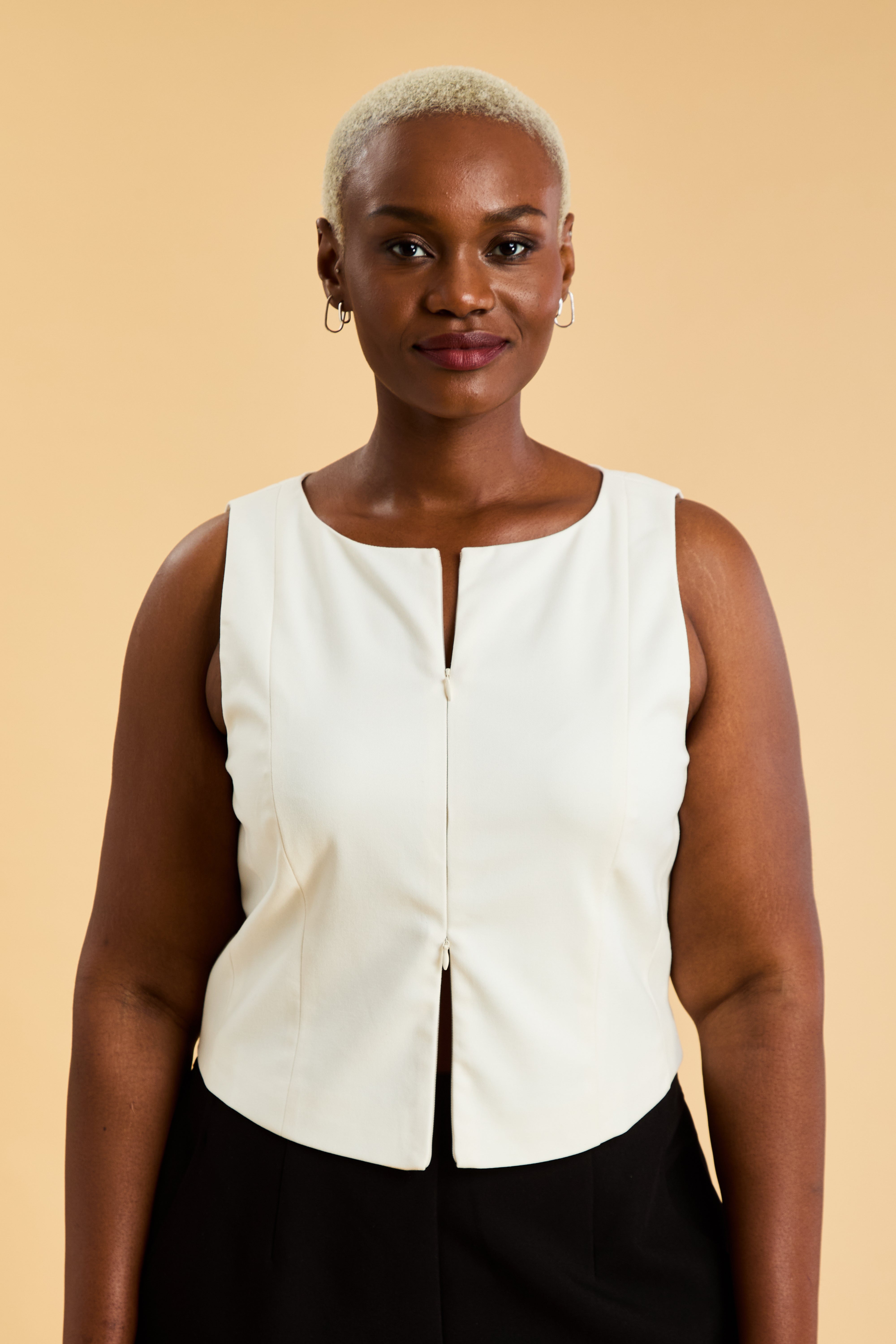 Woman wearing a White Triplicity sleeveless Alice double ended zipper top and Almond colored pants against a beige background close up front view