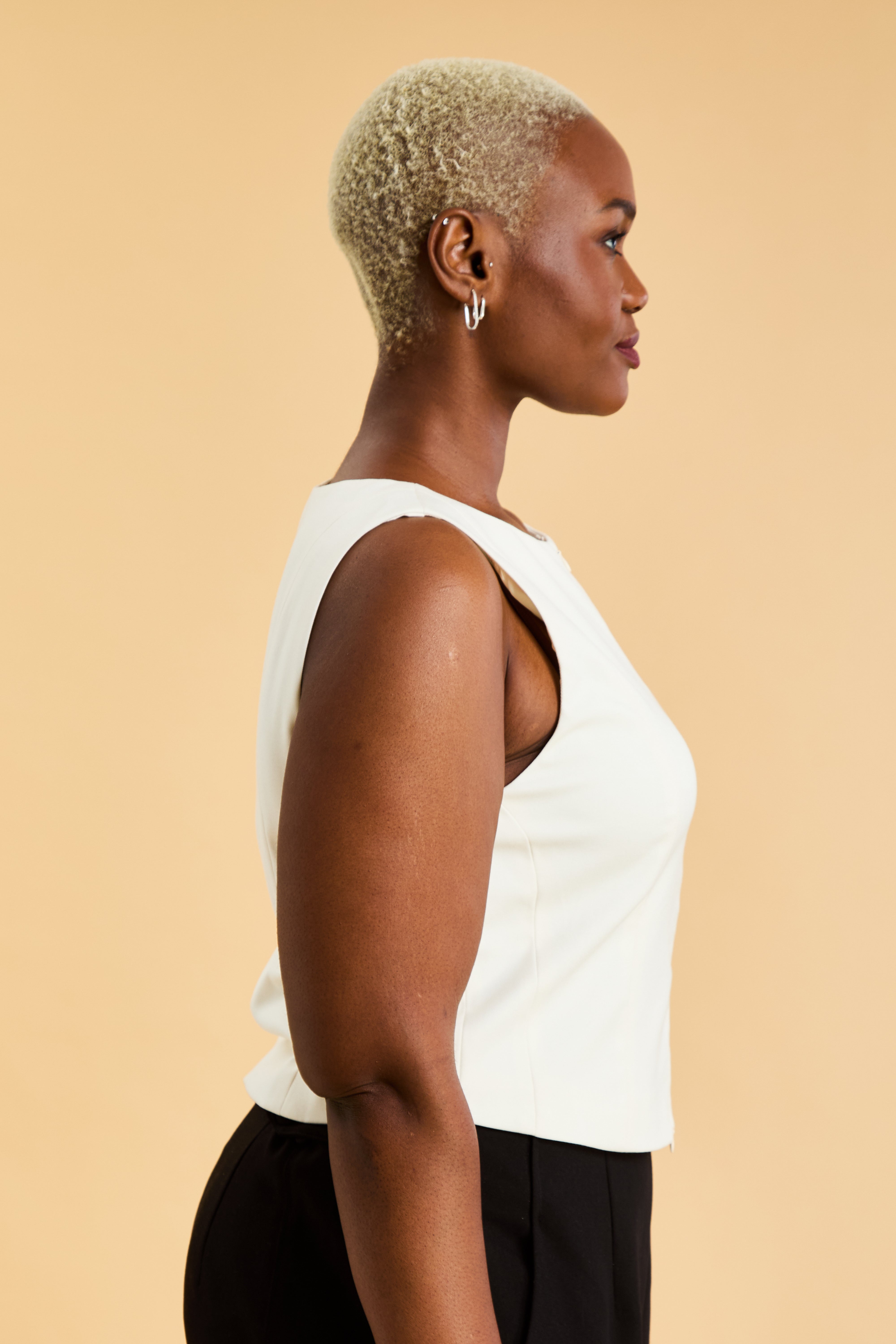 Woman wearing a White Triplicity sleeveless Alice double ended zipper top and Almond colored pants against a beige background close up side view