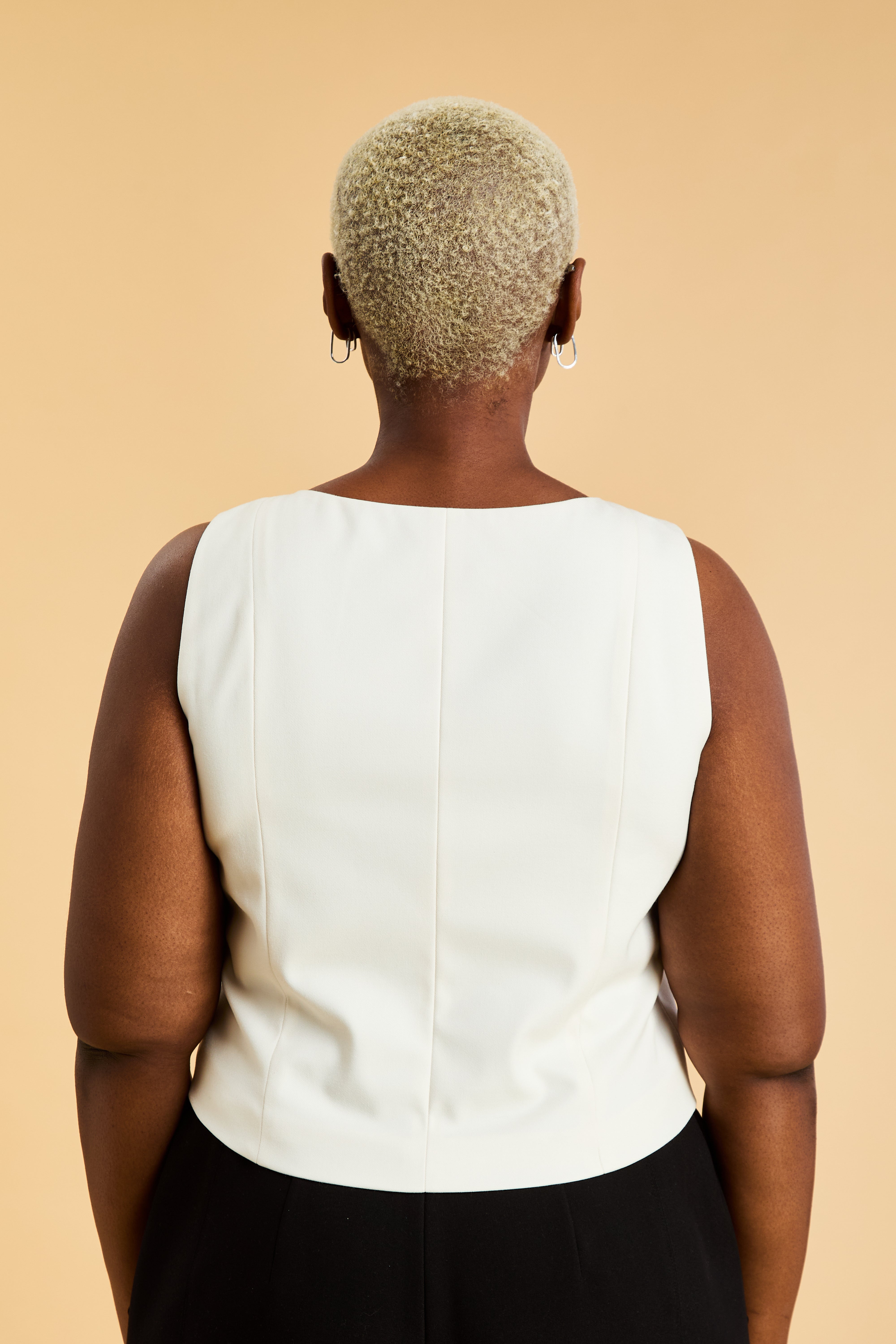 Woman wearing a White Triplicity sleeveless Alice double ended zipper top and Almond colored pants against a beige background close up back view