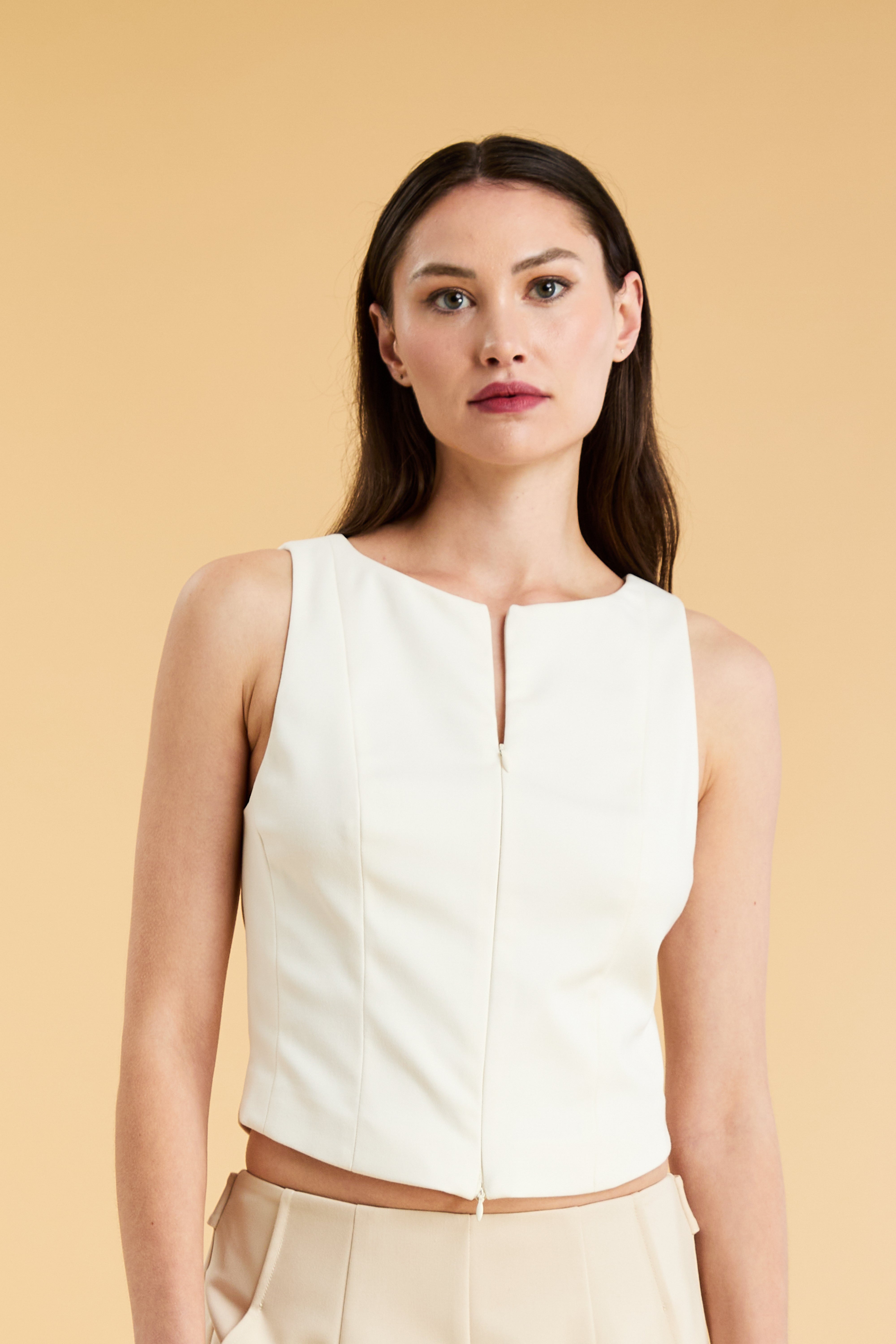 Woman wearing a White Triplicity sleeveless Alice double ended zipper top and Almond colored pants against a beige background close up front view