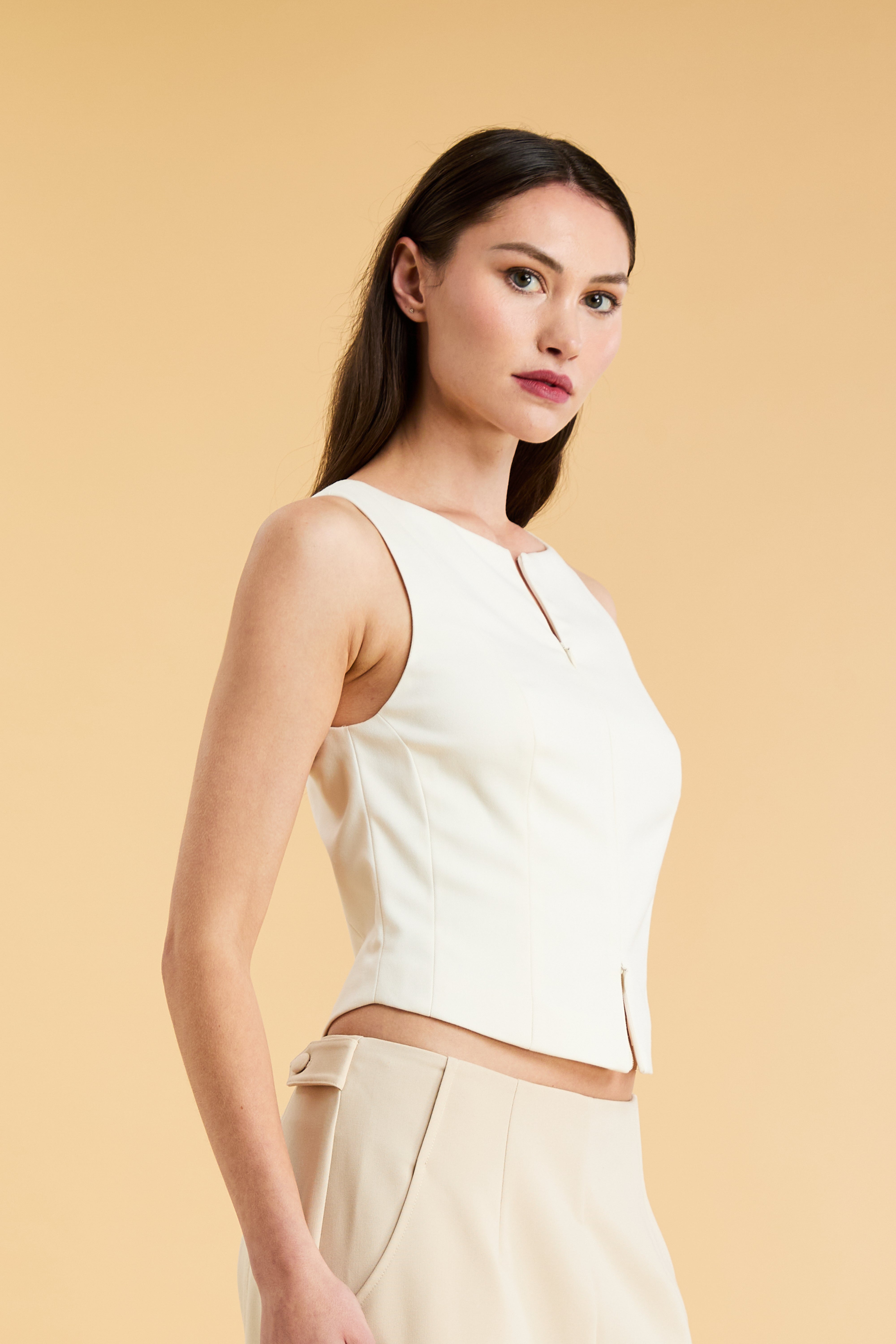 Woman wearing a White Triplicity sleeveless Alice double ended zipper top and Almond colored pants against a beige background close up three quarter front view