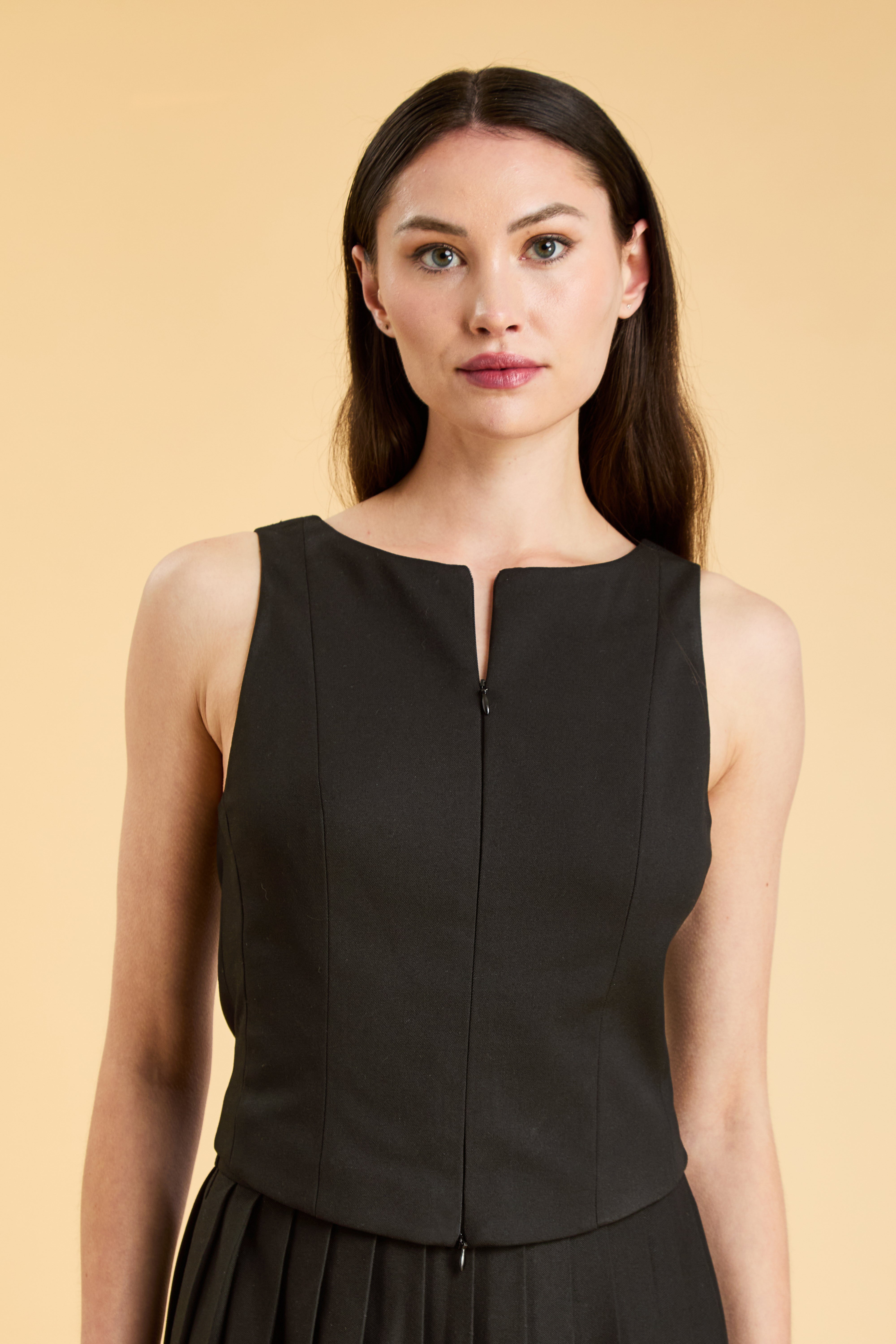 Person wearing a Black Alice double ended zipper Top on a Black pleated Elisabeth Mini skirt against a beige background