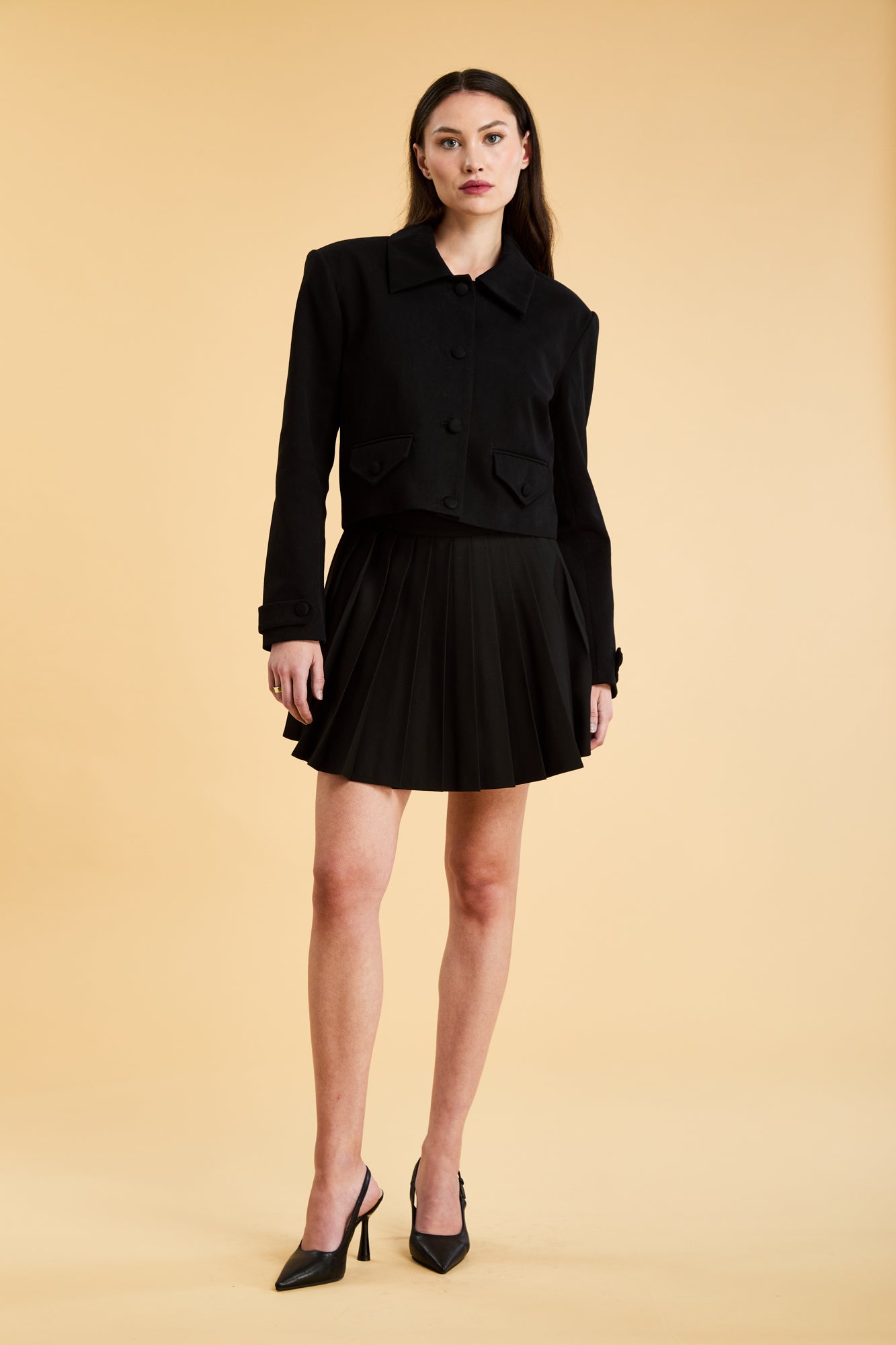 Woman wearing a Triplicity Charlotte Collared Cropped jacket and a Black Pleated Elisabeth mini skirt on a beige background