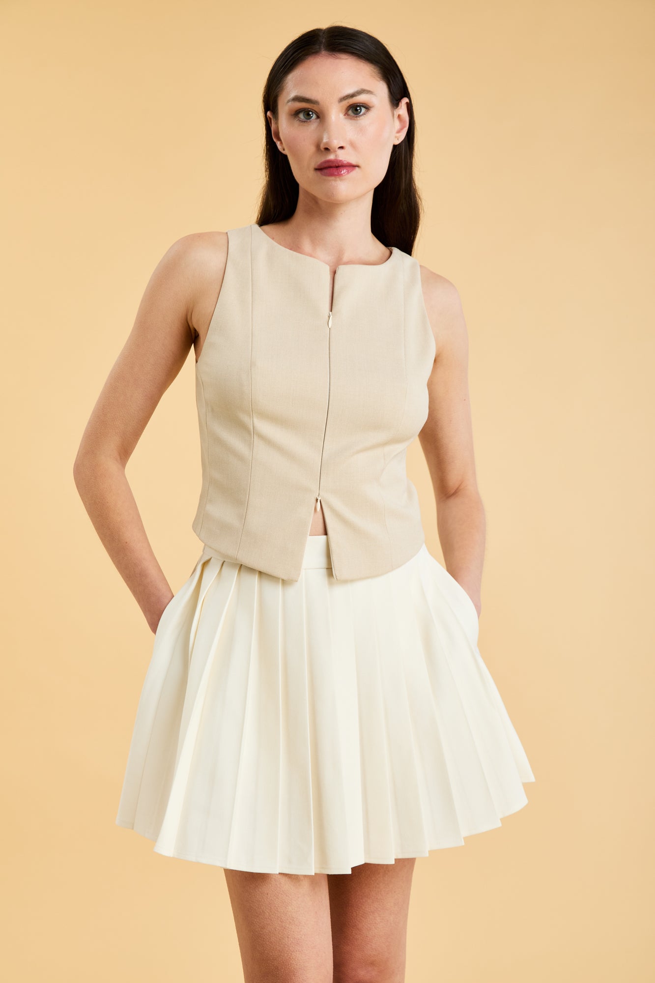 Woman wearing a Triplicity Alice Oat sleeveless double ended zipper top and Triplicity White pleated mini skirt against a beige background