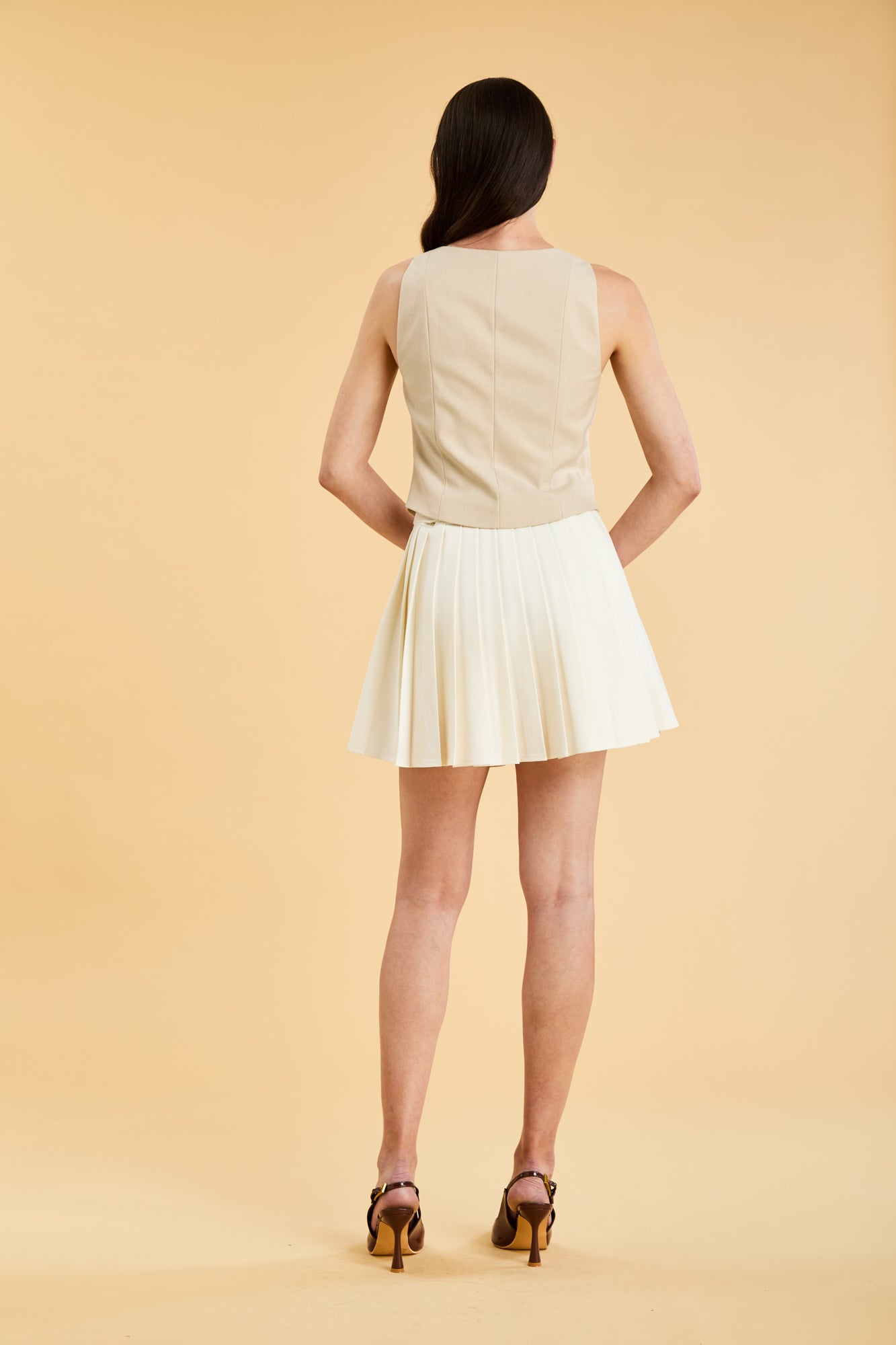 Woman wearing a Triplicity Alice Oat sleeveless double ended zipper top and Triplicity White pleated mini skirt against a beige background full body back view