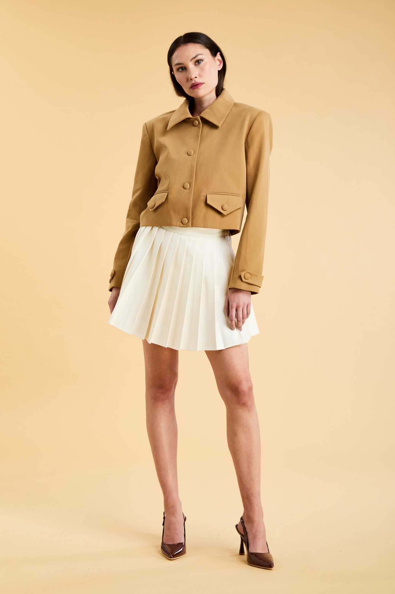 Woman wearing a Triplicity Camel colored Collared Cropped Charlotte jacket and Triplicity White pleated mini skirt against a beige background full body front view