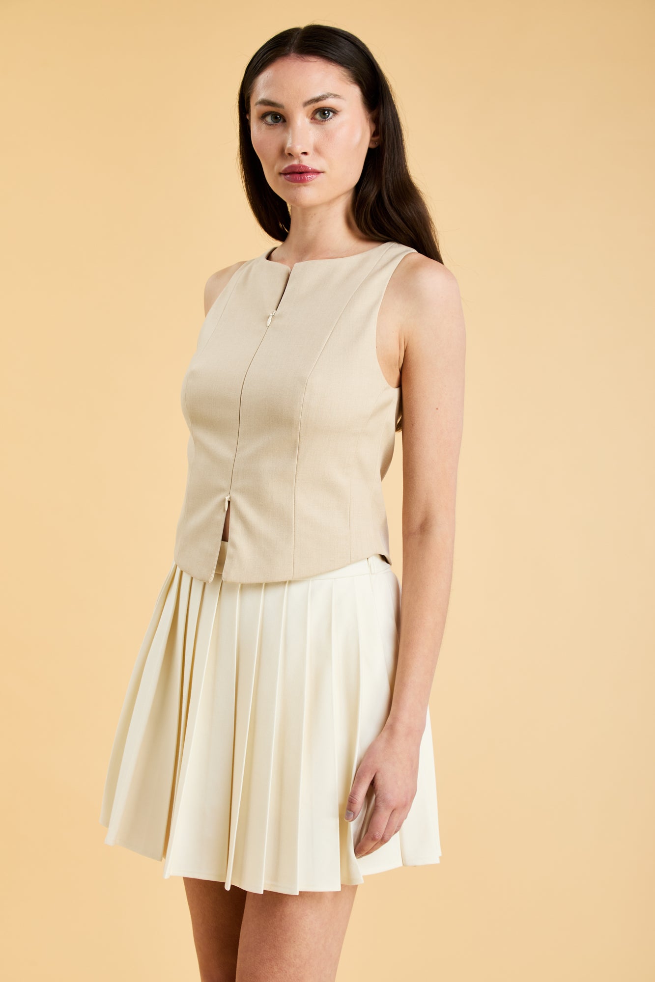 Woman wearing a Triplicity Alice Oat sleeveless double ended zipper top and Triplicity White pleated mini skirt against a beige background close up three quarter view