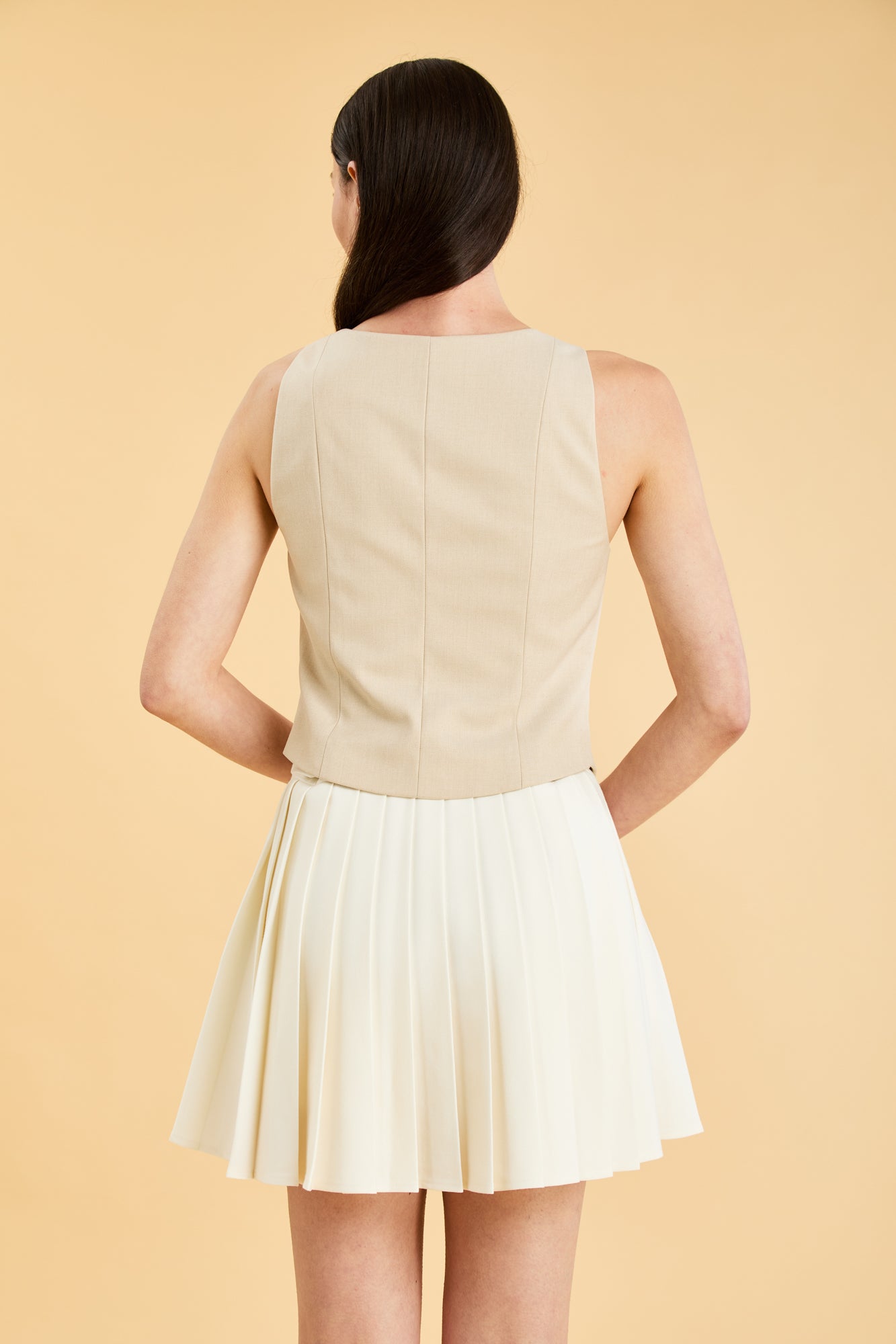 Woman wearing a Triplicity Alice Oat sleeveless double ended zipper top and Triplicity White pleated mini skirt against a beige background back view close up