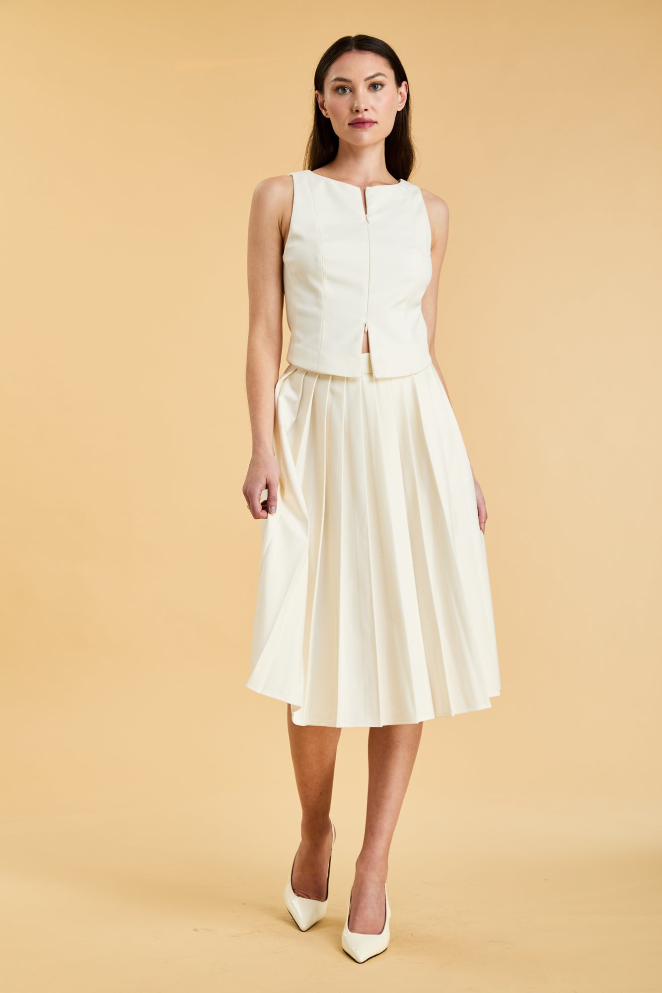Woman wearing a White colored Triplicity double ended zipper Alice top and White Triplicity pleated midi skirt against a beige background front view full body