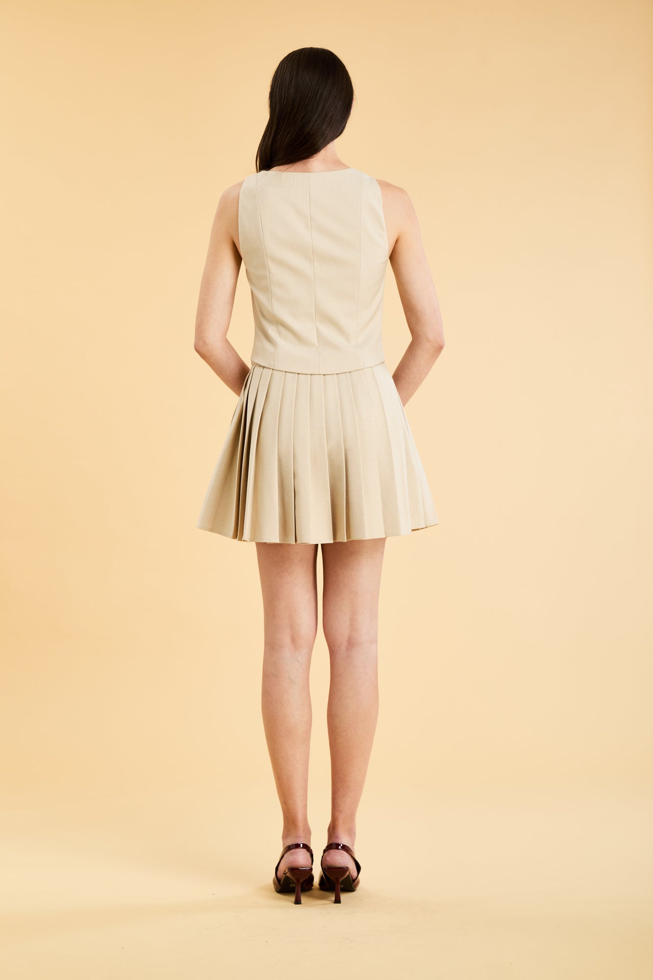 Woman wearing a Oat colored Triplicity Eloise Fitted Tank Top and a Oat Pleated Elisabeth mini skirt on a beige background full body back view