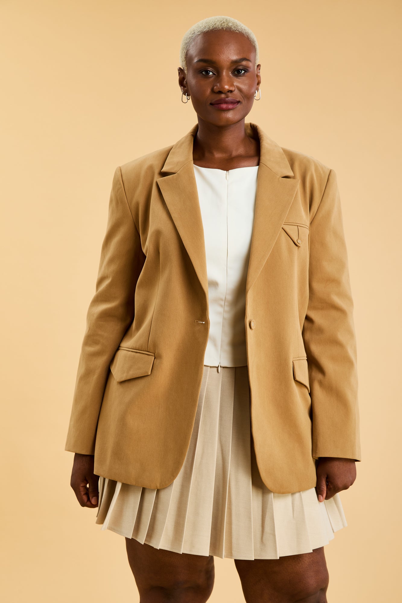 Woman wearing a Camel colored Constance Blazer and a Oat Pleated Elisabeth mini skirt on a beige background close up front view