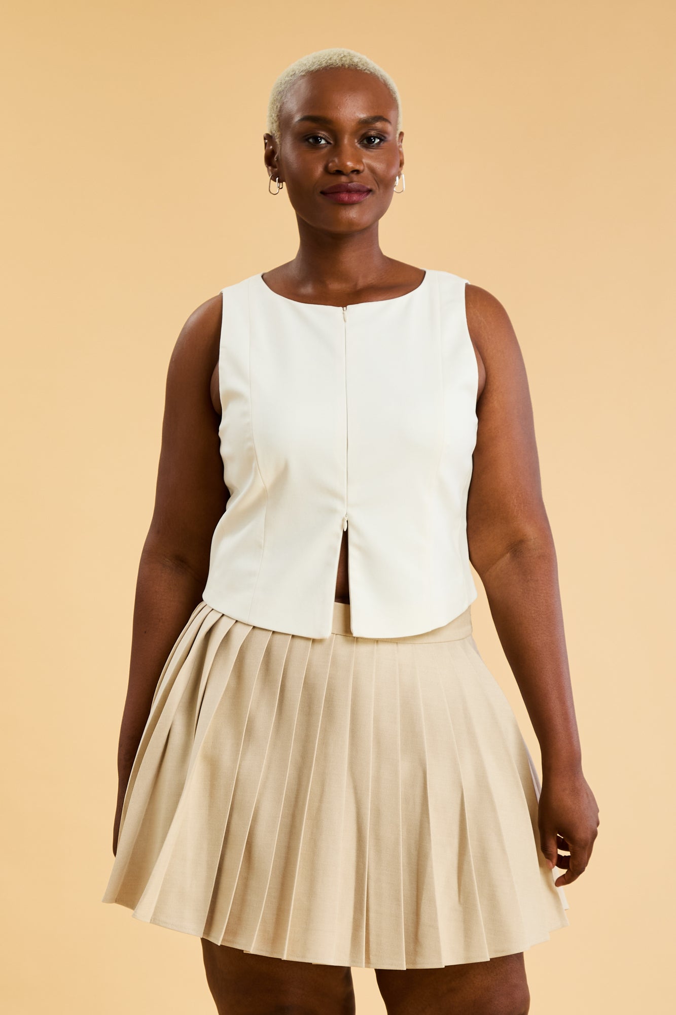 Woman wearing a White colored Triplicity Alice double ended zipper tank top and a Oat Pleated Elisabeth mini skirt on a beige background full body back view
