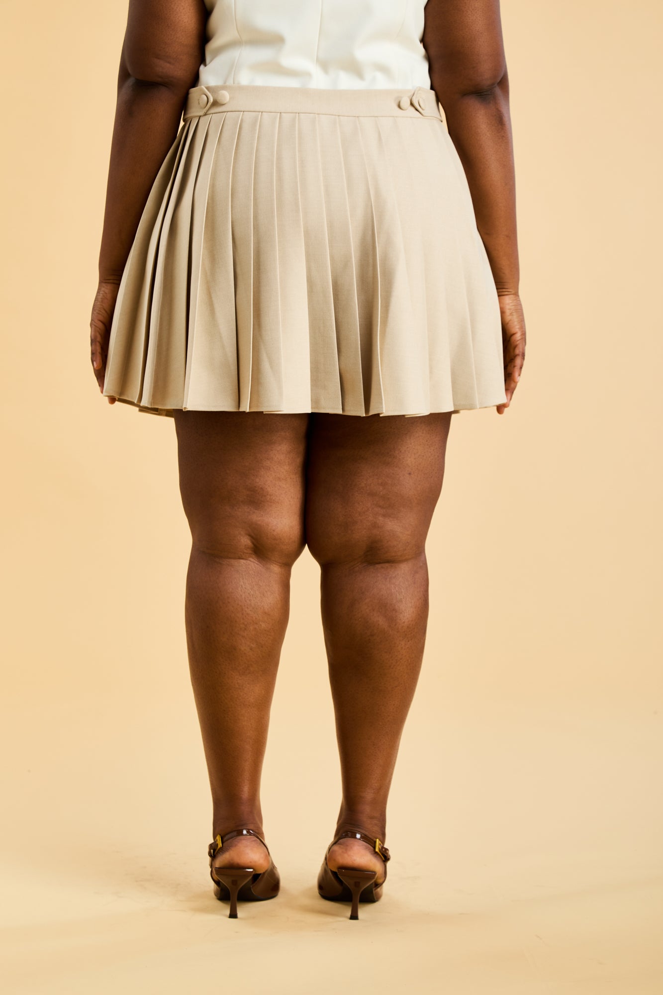 Woman wearing a White colored Triplicity Alice double ended zipper tank top and a Oat Pleated Elisabeth mini skirt on a beige background close up back view