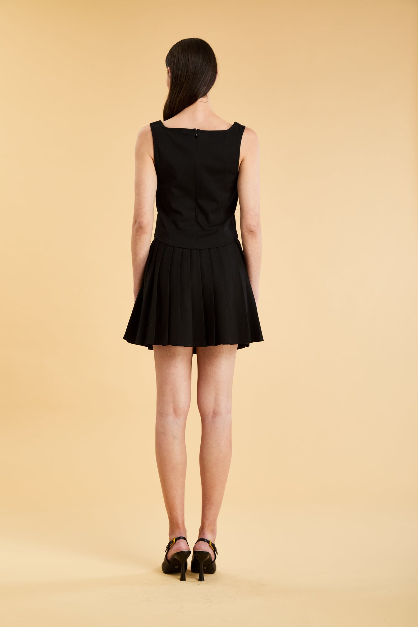 Woman wearing a Triplicity Eloise Fitted Tank Top and a Black Pleated Elisabeth mini skirt on a beige background full body back