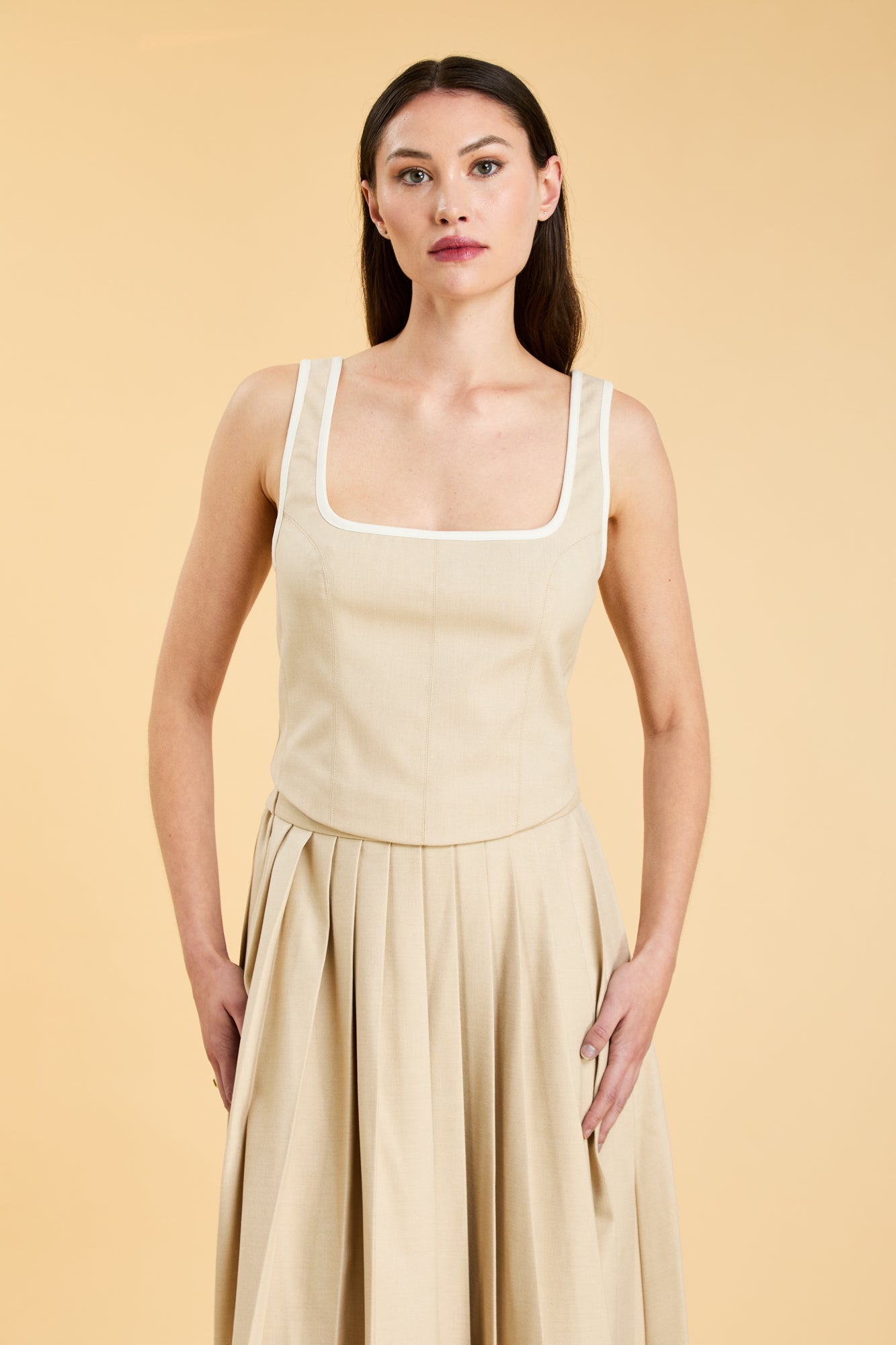 Woman wearing a Oat colored Triplicity Eloise Tank Top on a Oat colored Elisabeth pleated midi skirt against a beige background side view close up