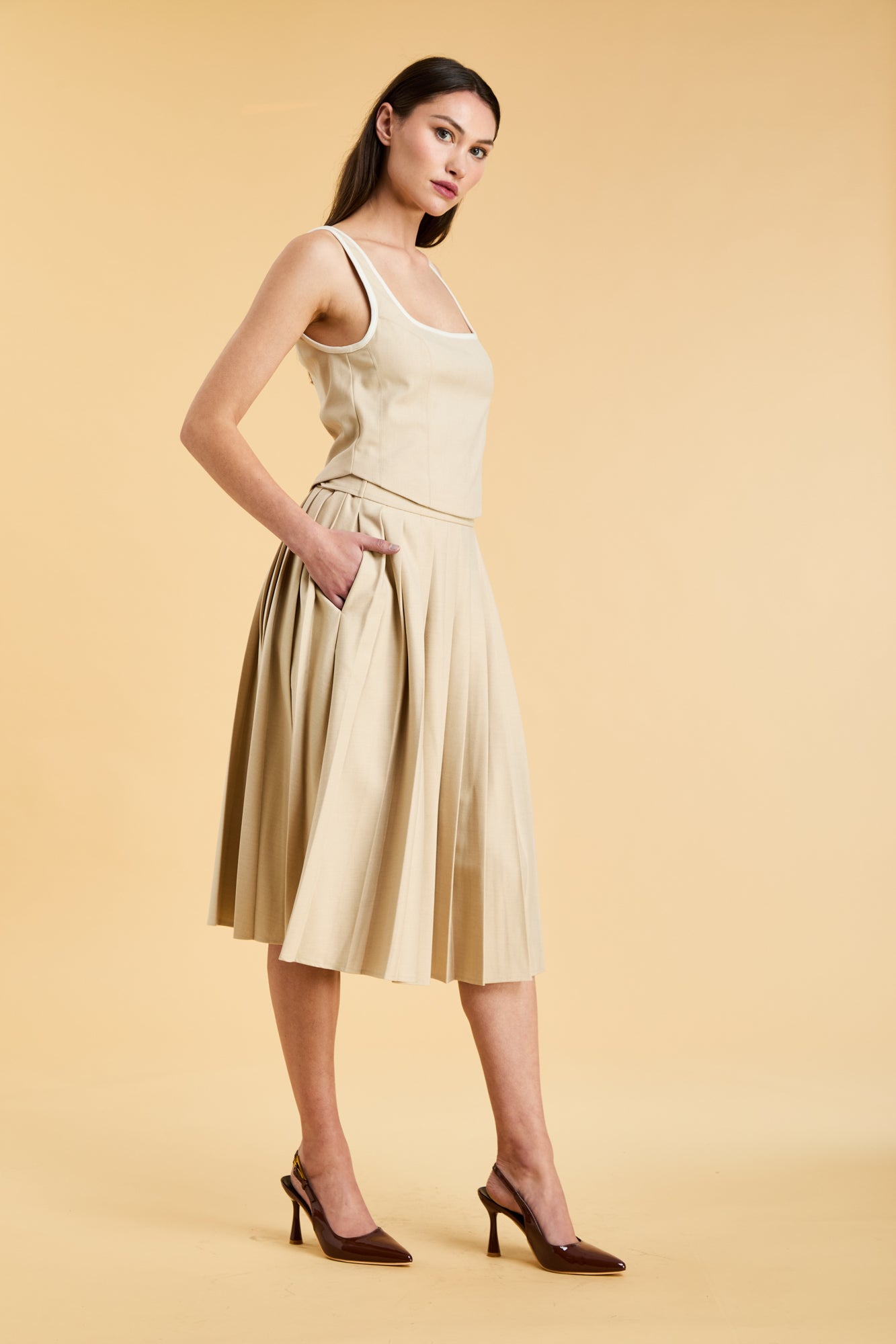 Close up of a woman wearing a Triplicity Oat Elisabeth pleated mini skirt against a beige background three quarter view