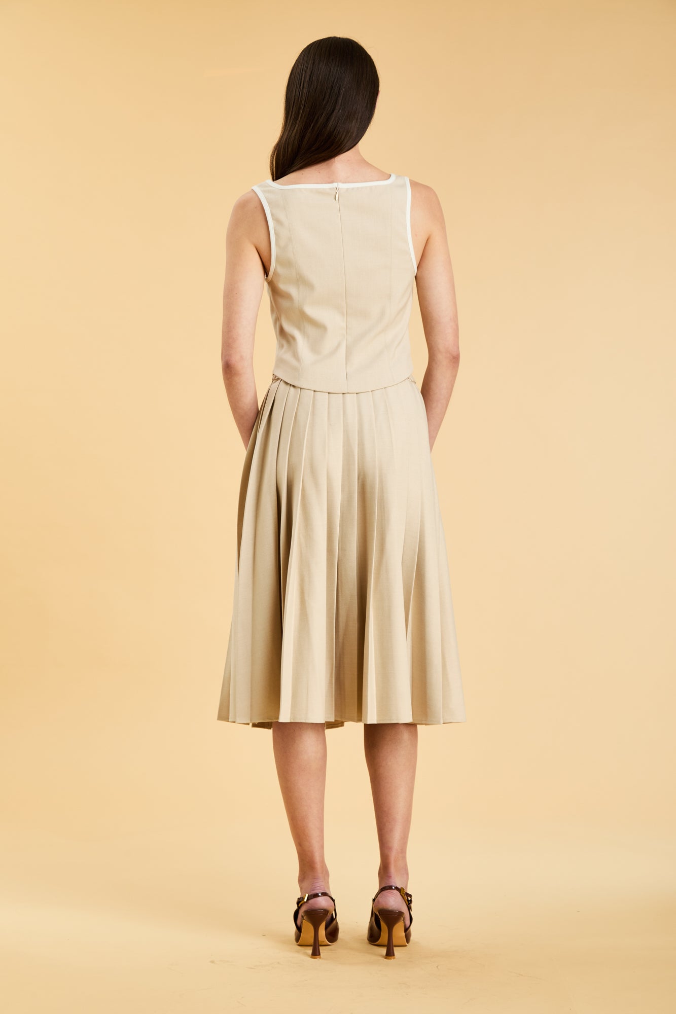 Close up of a woman wearing a Triplicity Oat Elisabeth pleated mini skirt against a beige background back view
