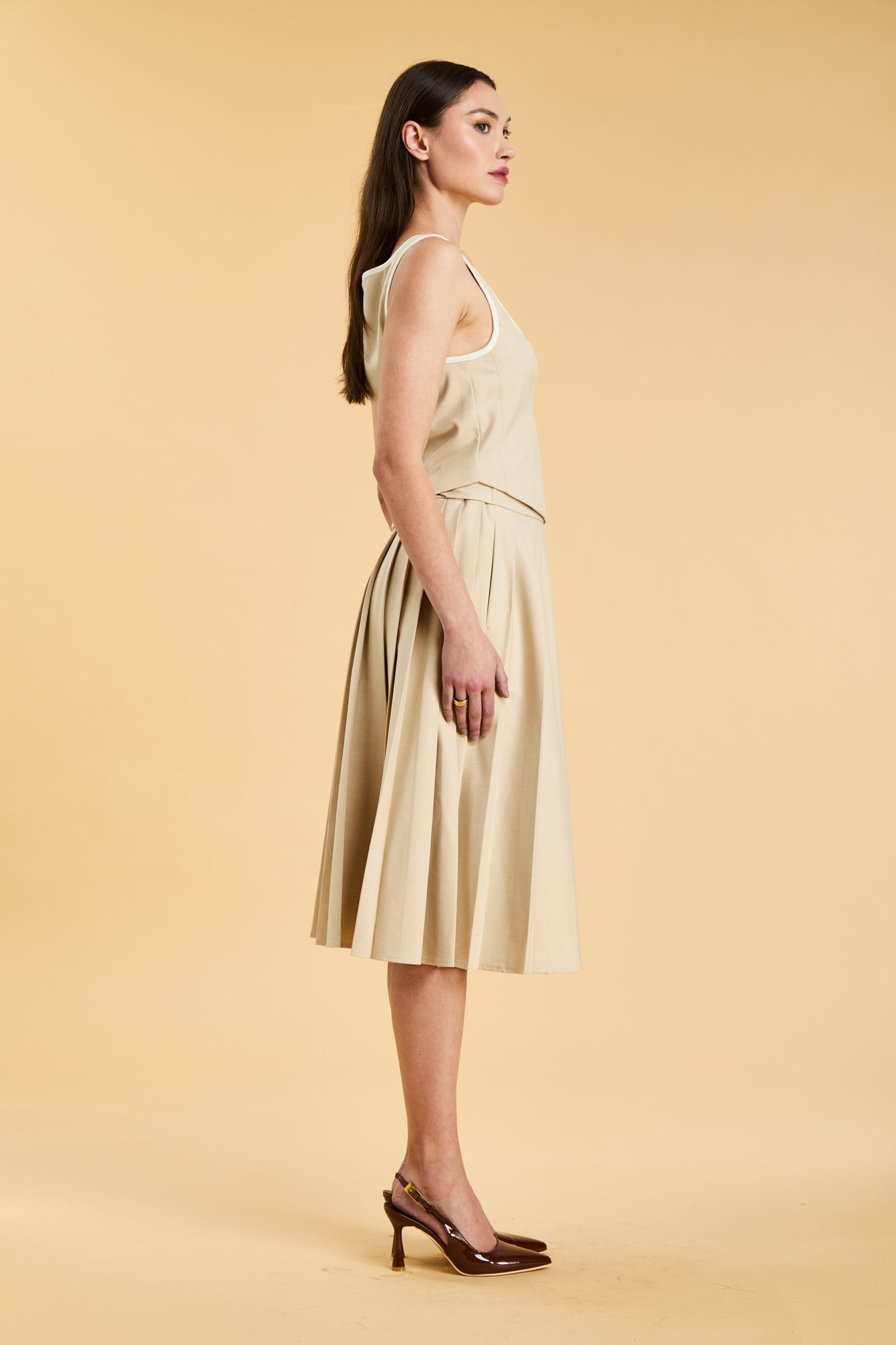 Close up of a woman wearing a Triplicity Oat Elisabeth pleated mini skirt against a beige background side view
