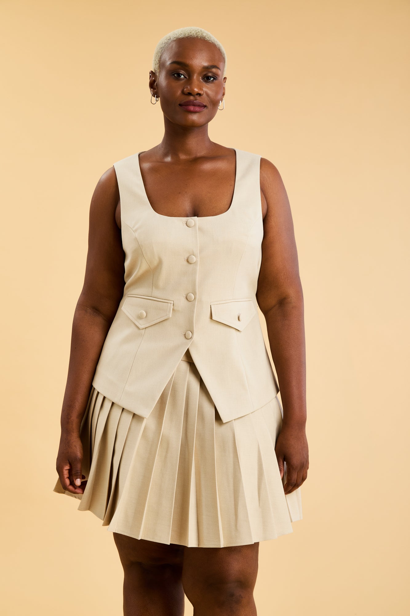 Woman wearing an Oat colored Triplicity sleeveless Georgiana vest over a White pleated midi skirt on a beige background front view close up