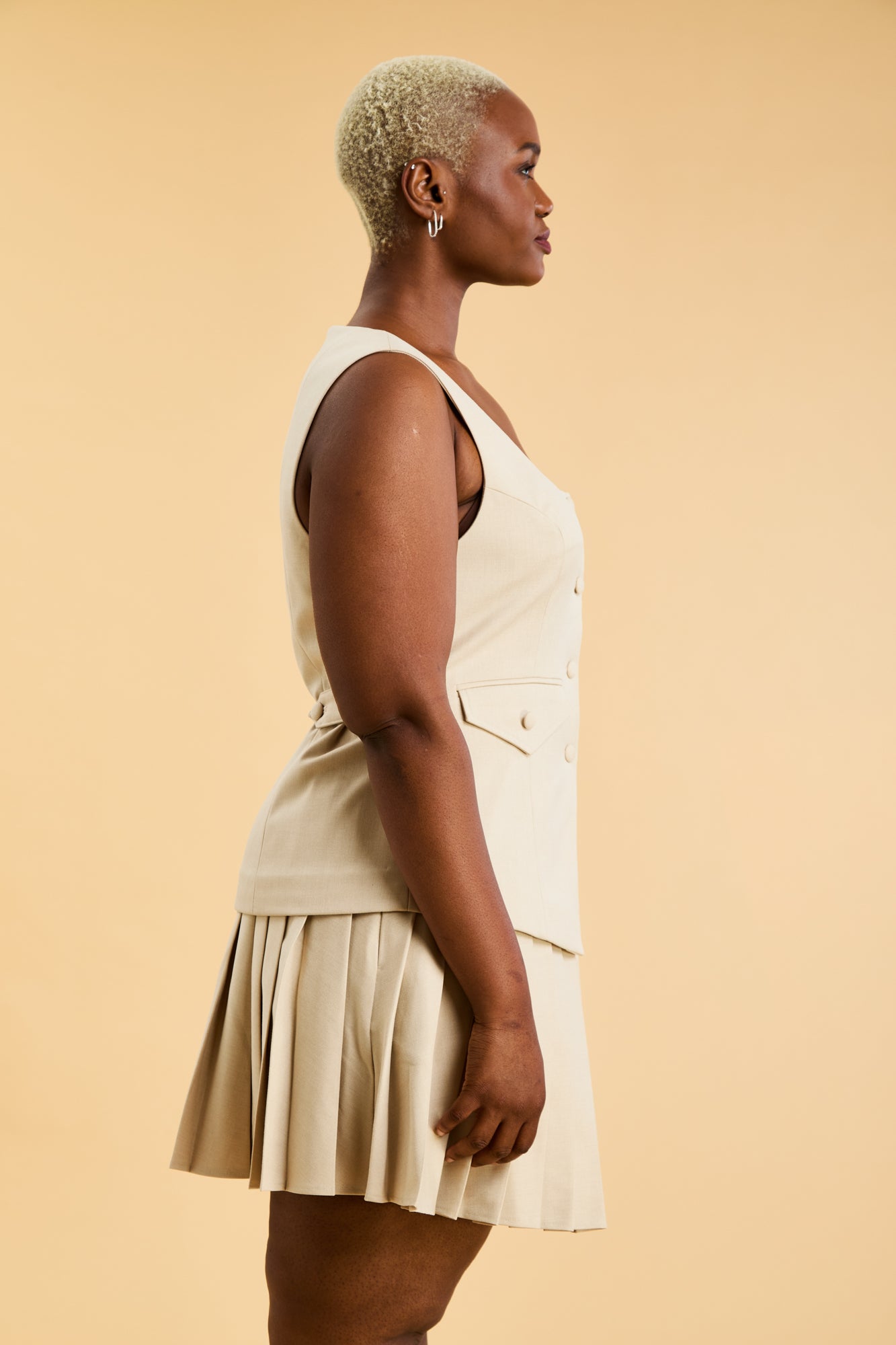 Woman wearing an Oat colored Triplicity sleeveless Georgiana vest over a White pleated midi skirt on a beige background side view close up