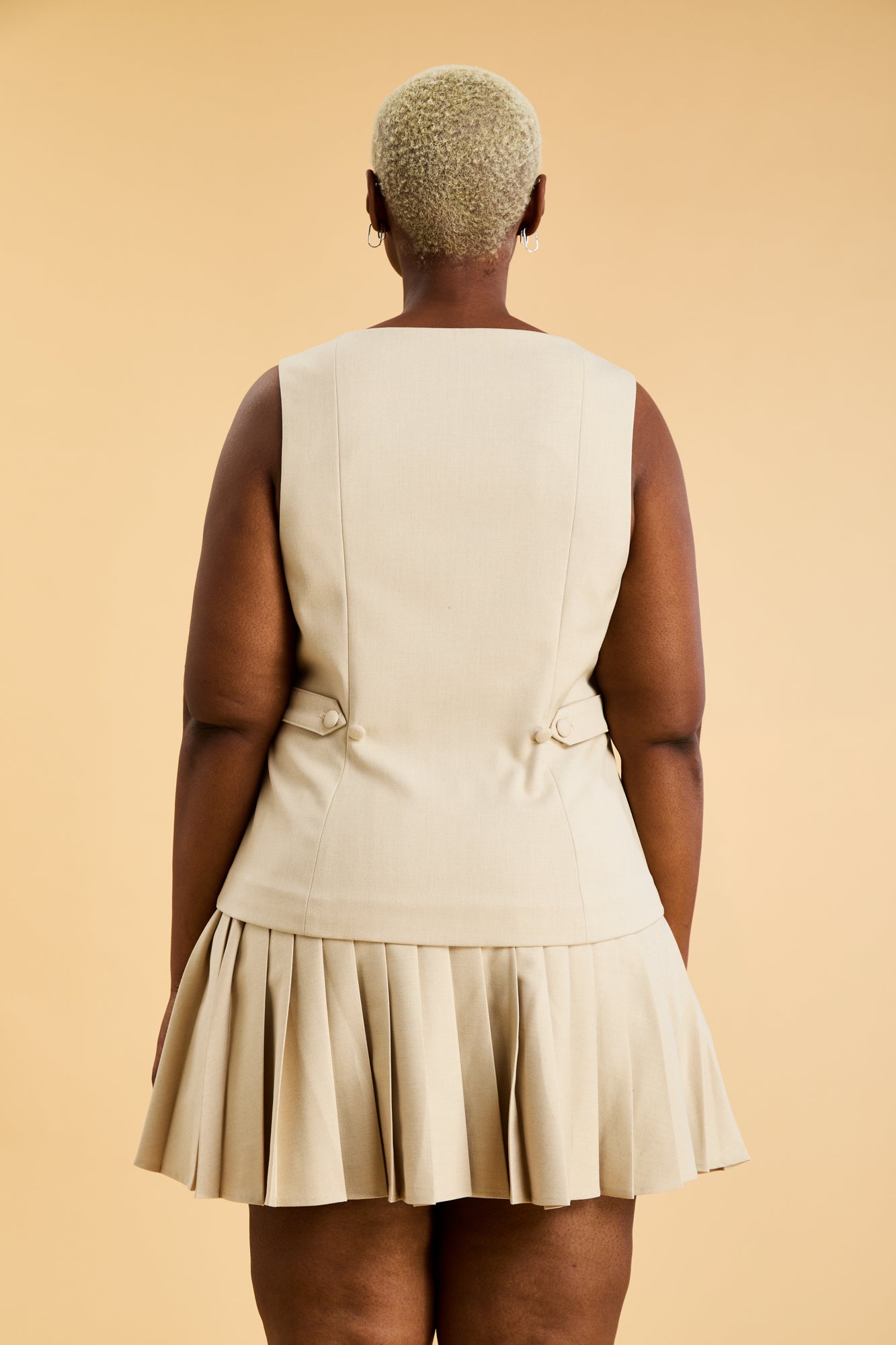 Woman wearing an Oat colored Triplicity sleeveless Georgiana vest over a White pleated midi skirt on a beige background front view close up