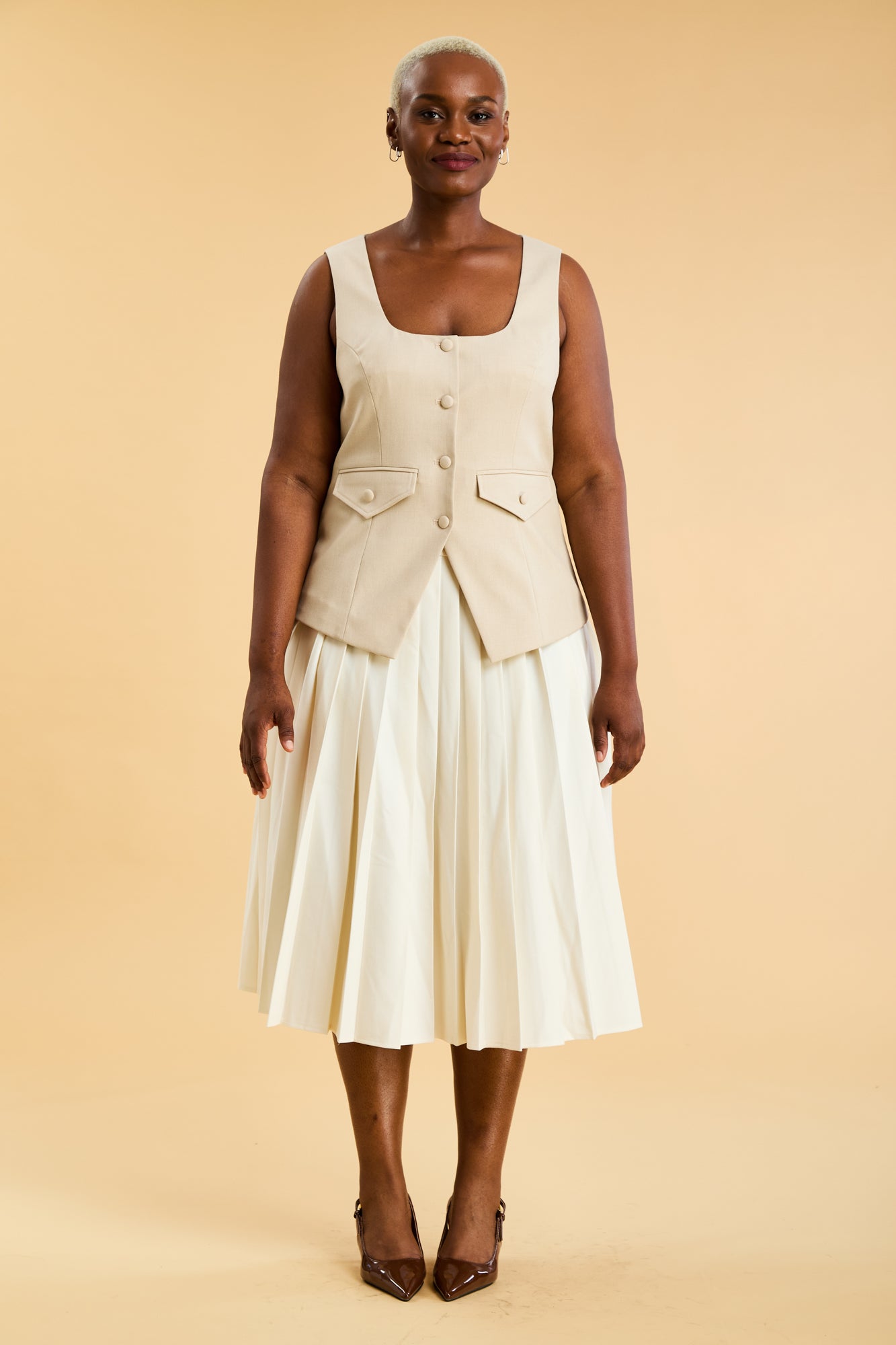 Woman wearing an Oat colored Triplicity sleeveless Georgiana vest over a White pleated midi skirt on a beige background front view full body