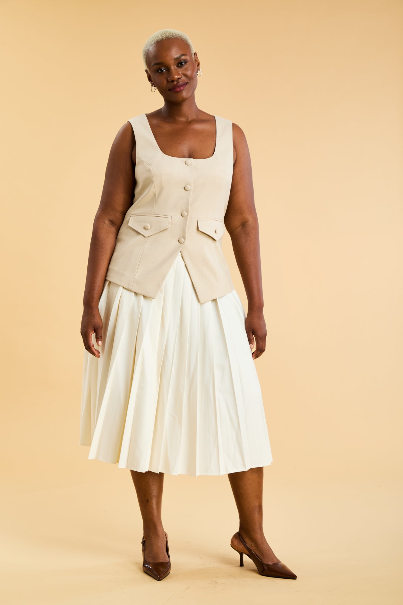 Woman wearing an Oat colored Triplicity sleeveless Georgiana vest over a White pleated midi skirt on a beige background