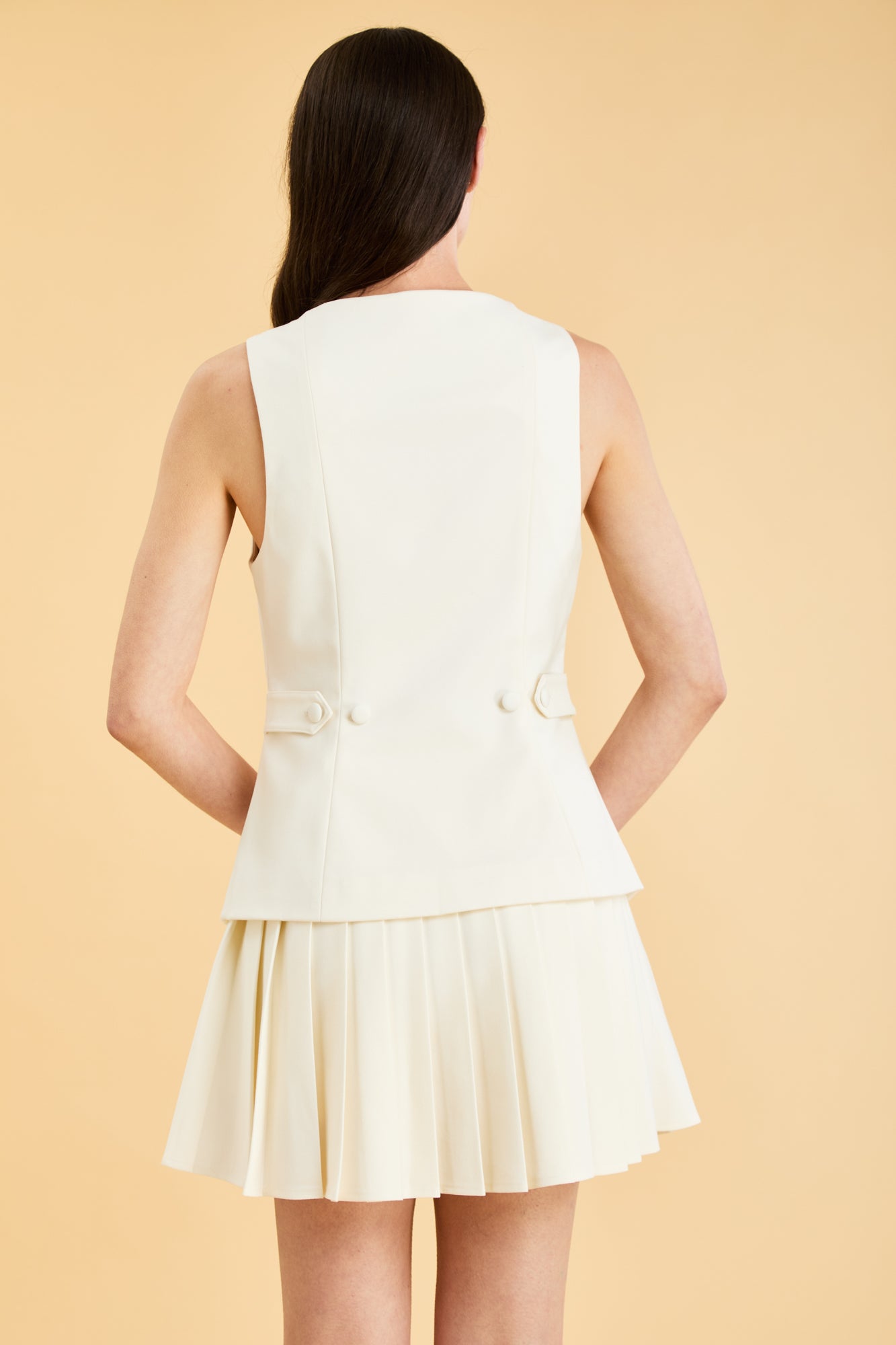Woman wearing a White Triplicity Georgiana vest top on a Triplicity White pleated midi skirt against a beige background back view close up