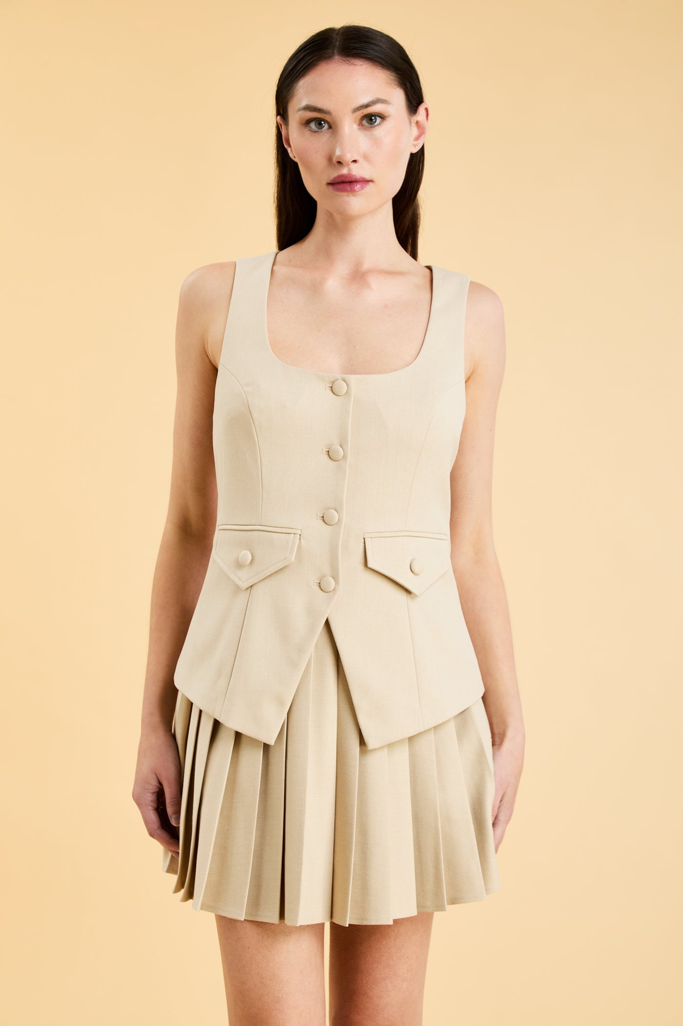 Woman wearing an Oat colored Triplicity sleeveless Georgiana vest over a White pleated midi skirt on a beige background front view close up