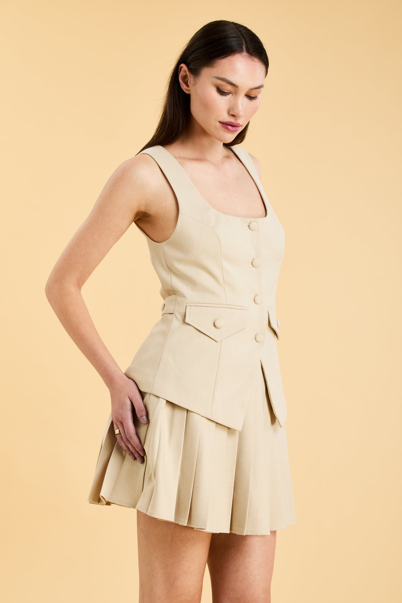 Woman wearing an Oat colored Triplicity sleeveless Georgiana vest over a White pleated midi skirt on a beige background three quarter view close up