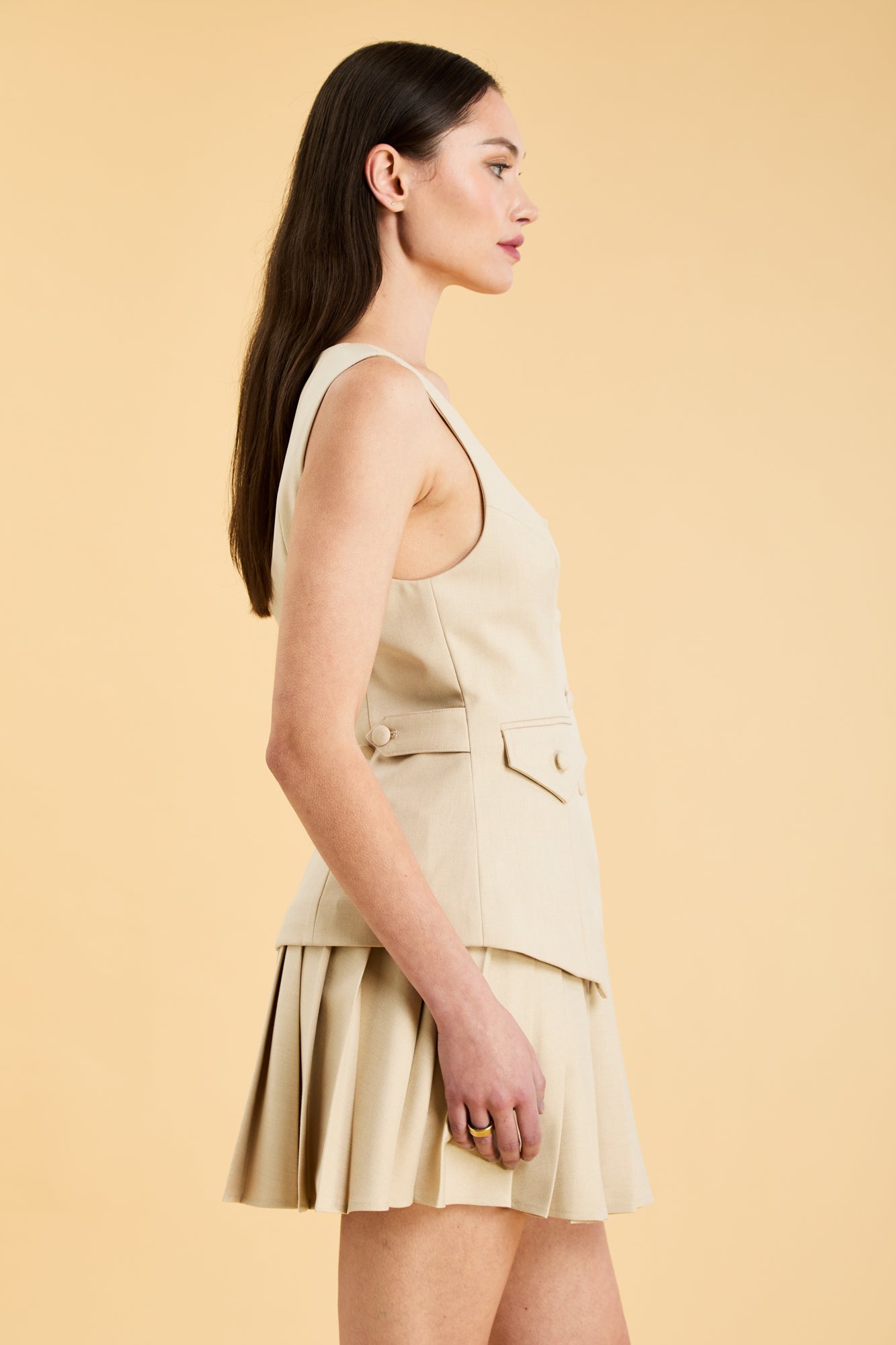 Woman wearing an Oat colored Triplicity sleeveless Georgiana vest over a White pleated midi skirt on a beige background side view close up