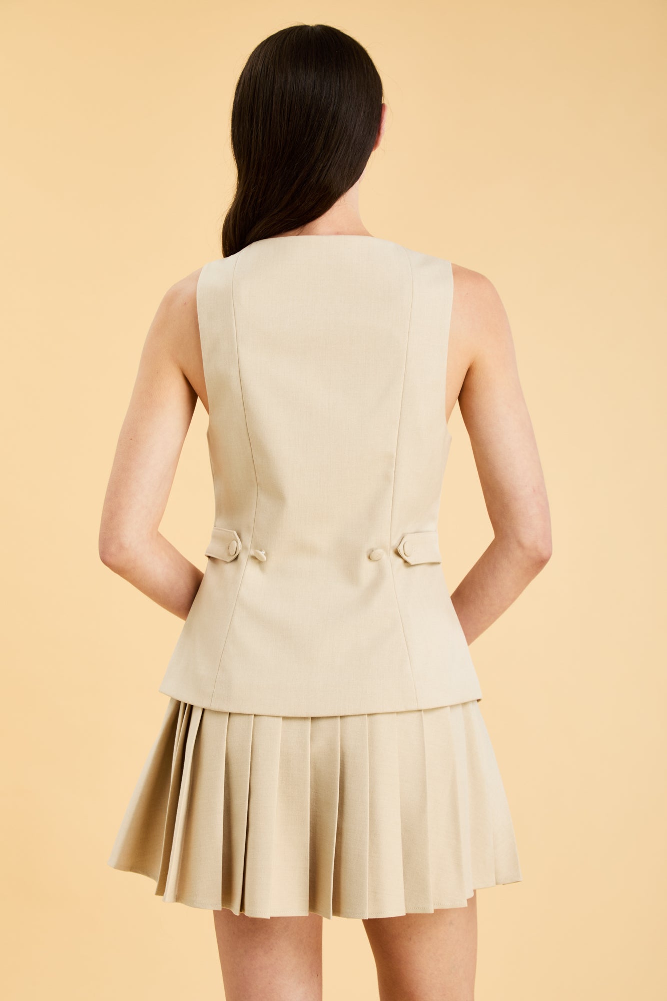 Woman wearing an Oat colored Triplicity sleeveless Georgiana vest over a White pleated midi skirt on a beige background front view close up