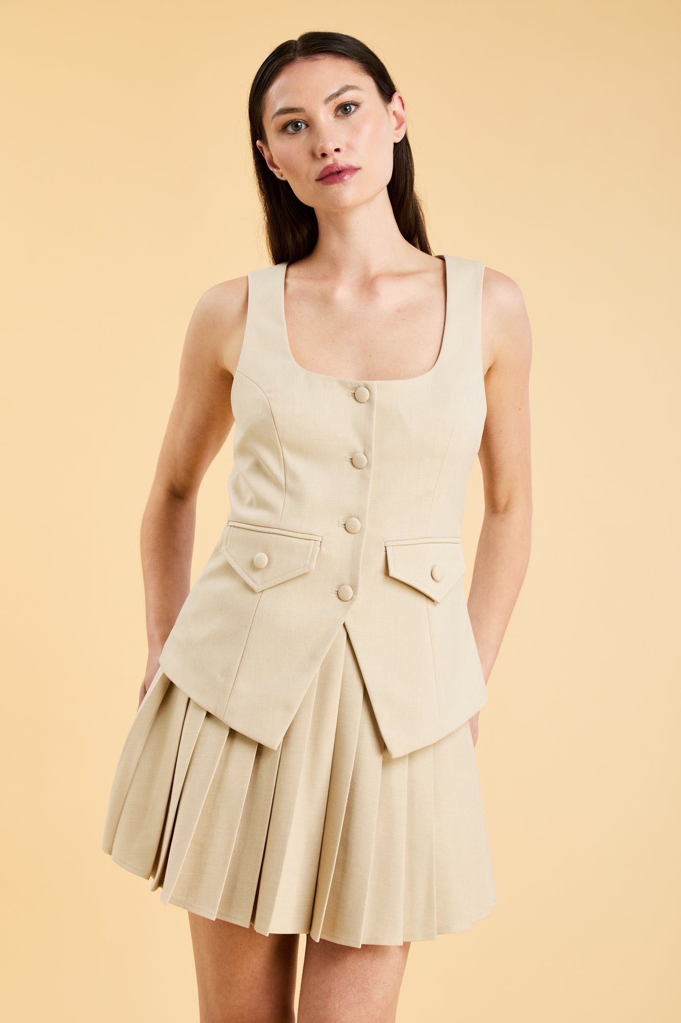 Woman wearing an Oat colored Triplicity sleeveless Georgiana vest over a White pleated midi skirt on a beige background front view close up