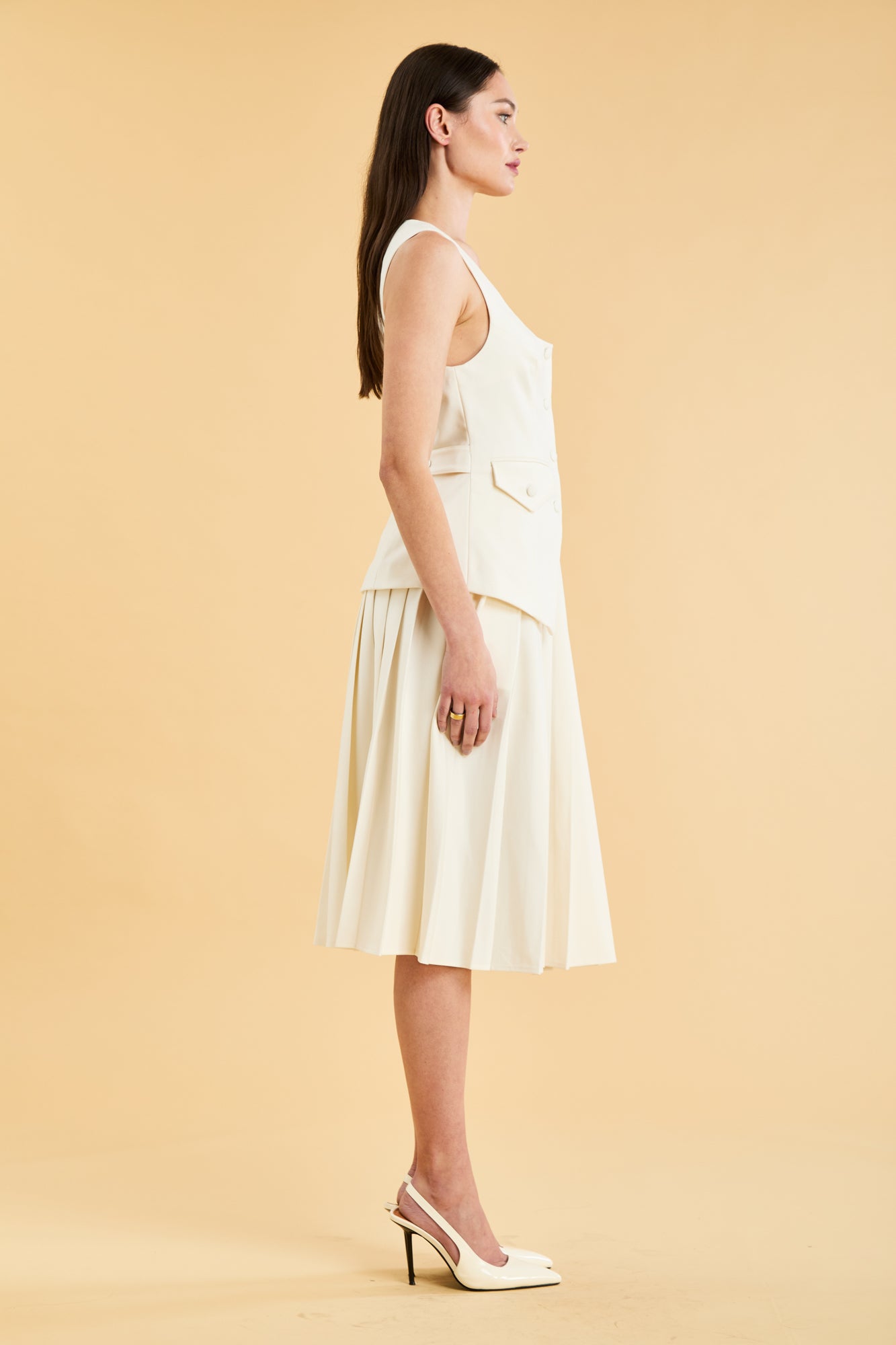 Woman wearing a White Triplicity Georgiana vest top on a Triplicity White pleated midi skirt against a beige background side view full body