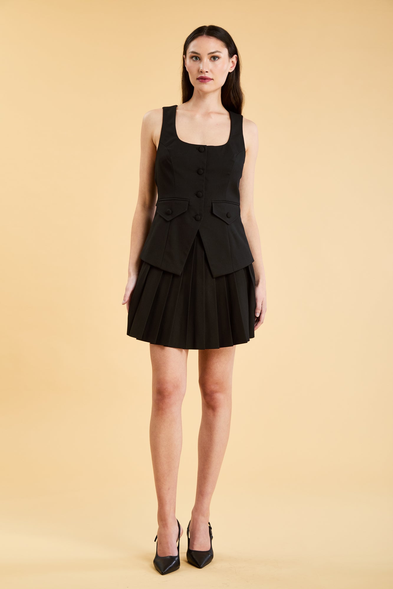 Woman wearing a Black Triplicity Georgiana Vest top on the Triplicity Elisabeth pleated mini skirt on a beige background