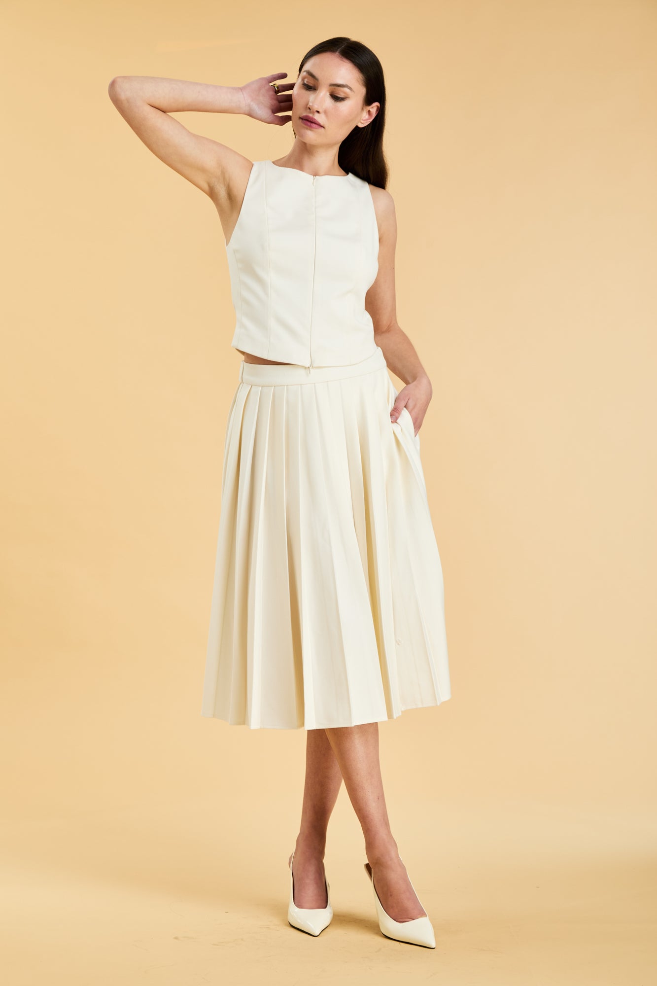 Woman wearing a White colored Triplicity double ended zipper Alice top and White Triplicity pleated midi skirt against a beige background front view full body