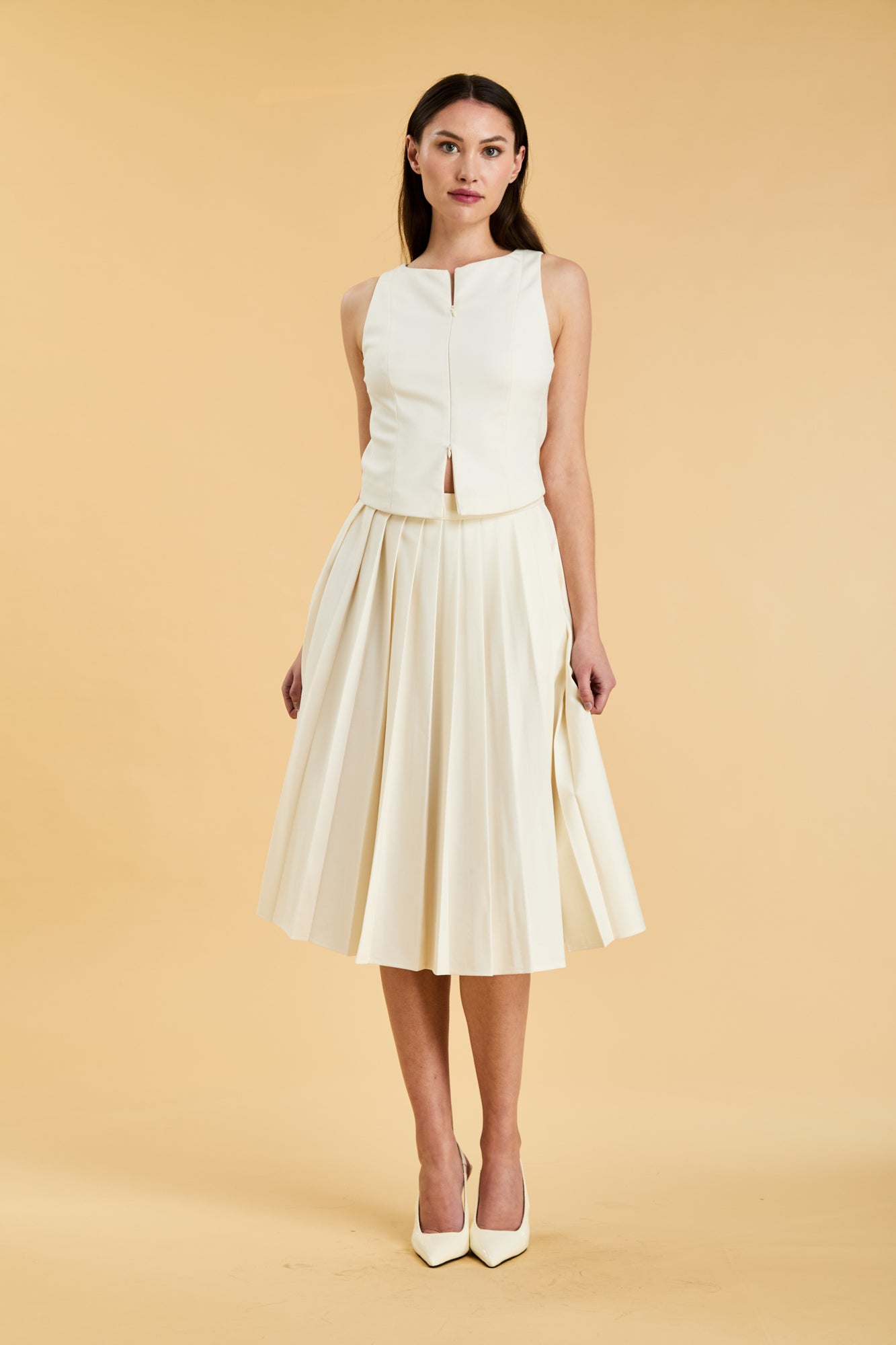 Woman wearing a White colored Triplicity double ended zipper Alice top and White Triplicity pleated midi skirt against a beige background front view full body