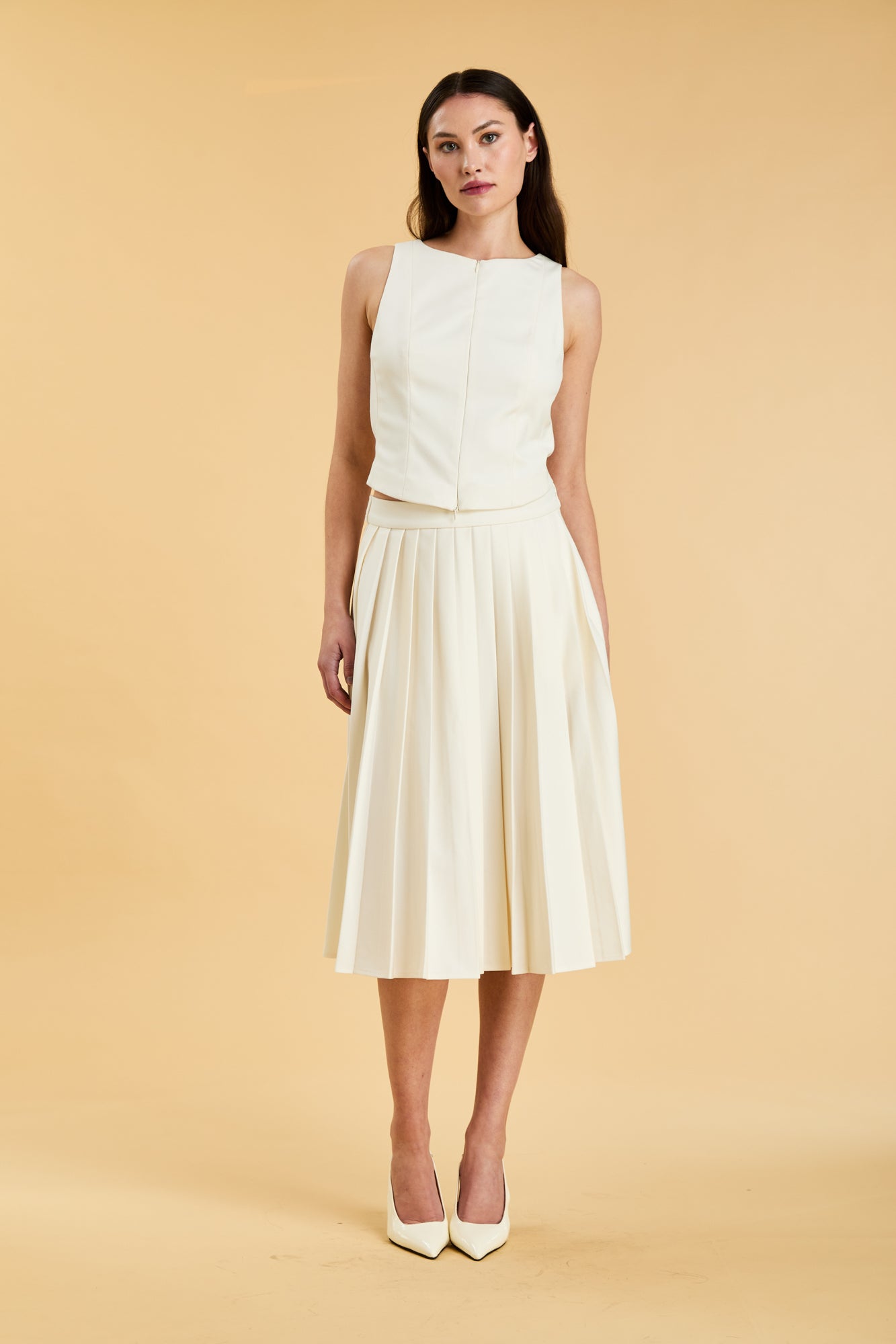 Woman wearing a White colored Triplicity double ended zipper Alice top and White Triplicity pleated midi skirt against a beige background front view full body