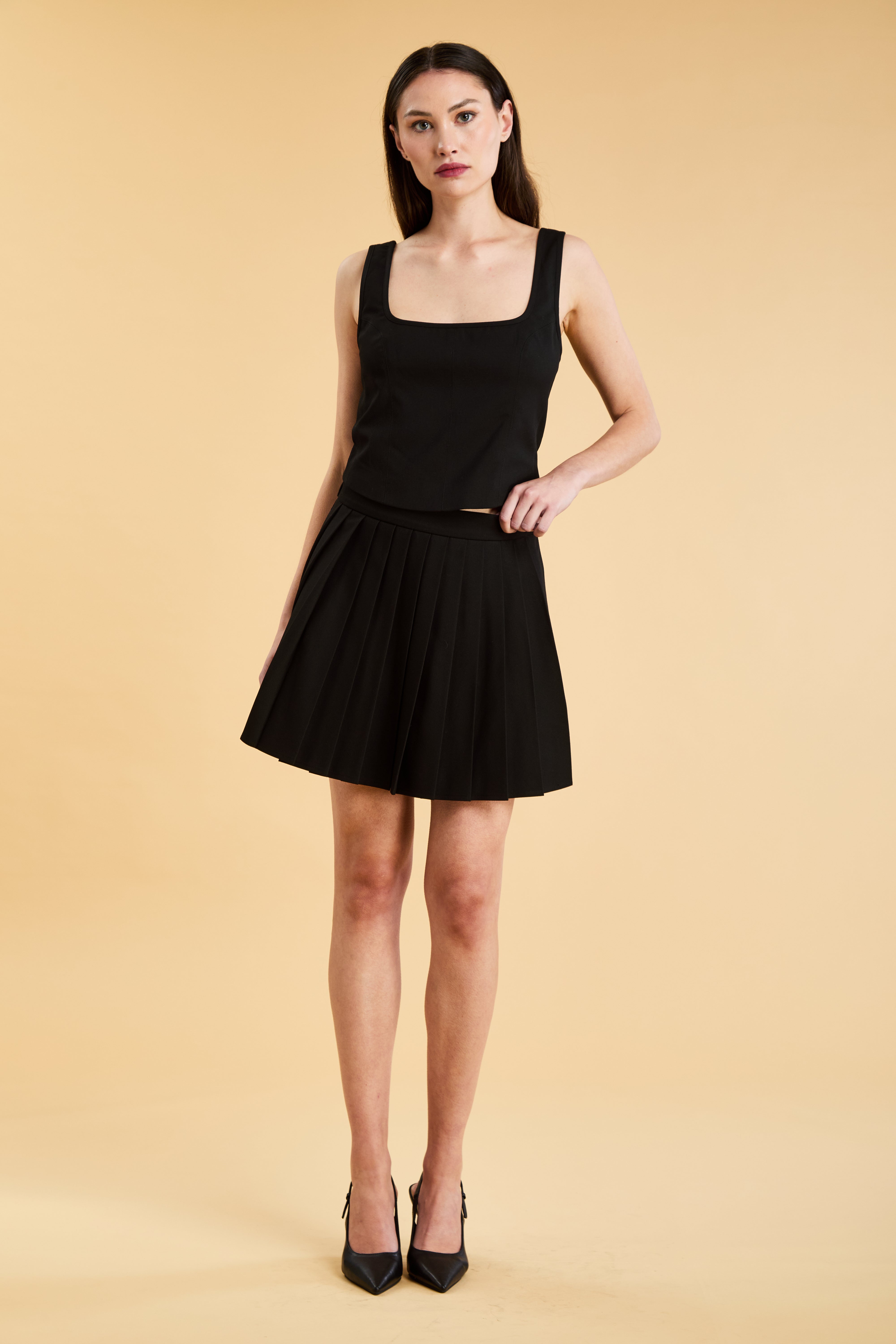Woman wearing a Triplicity Eloise Fitted Tank Top and a Black Pleated Elisabeth mini skirt on a beige background full body front view