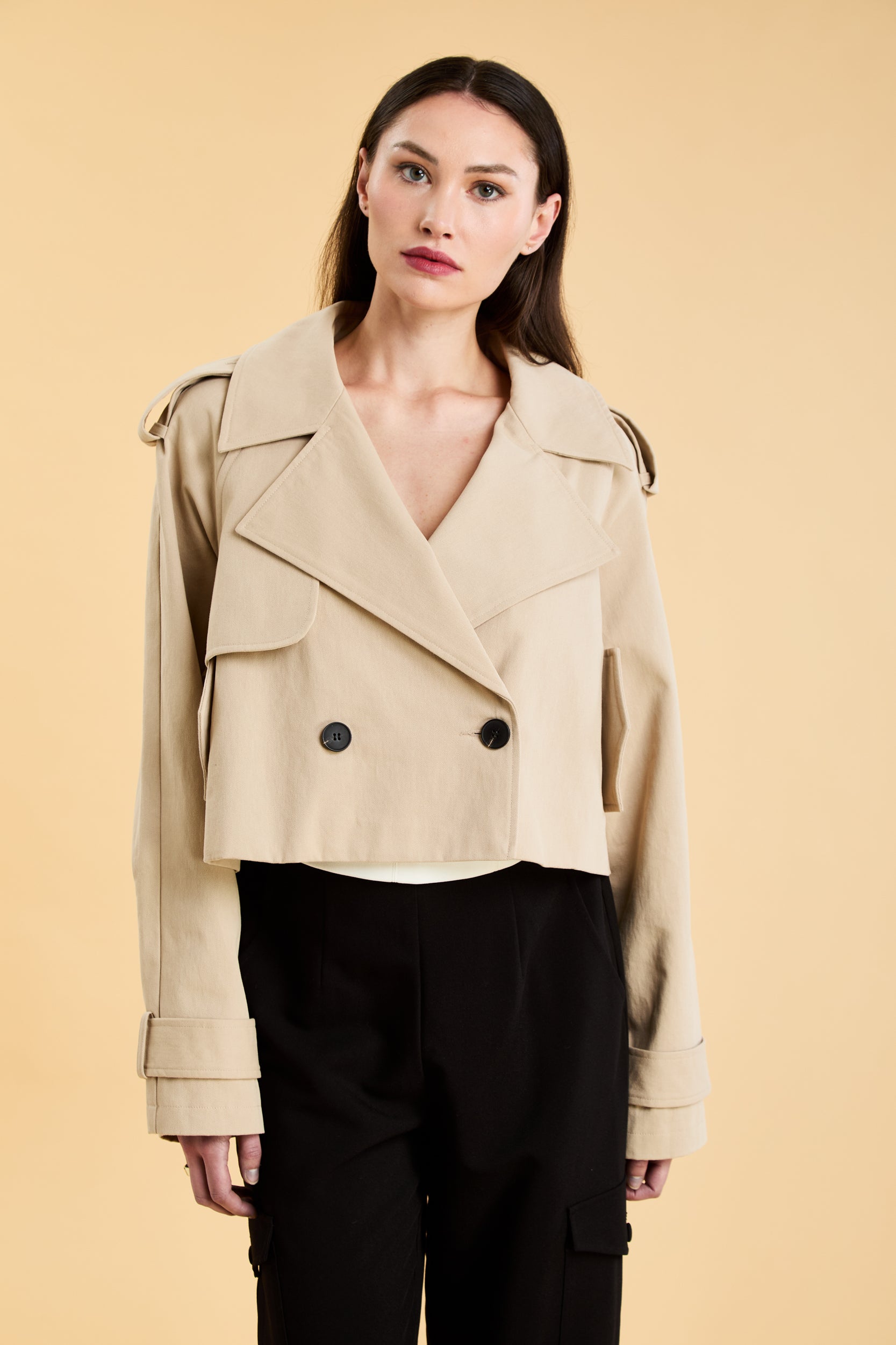 Woman wearing a beige cropped trench coat jacket in stone color against a beige background