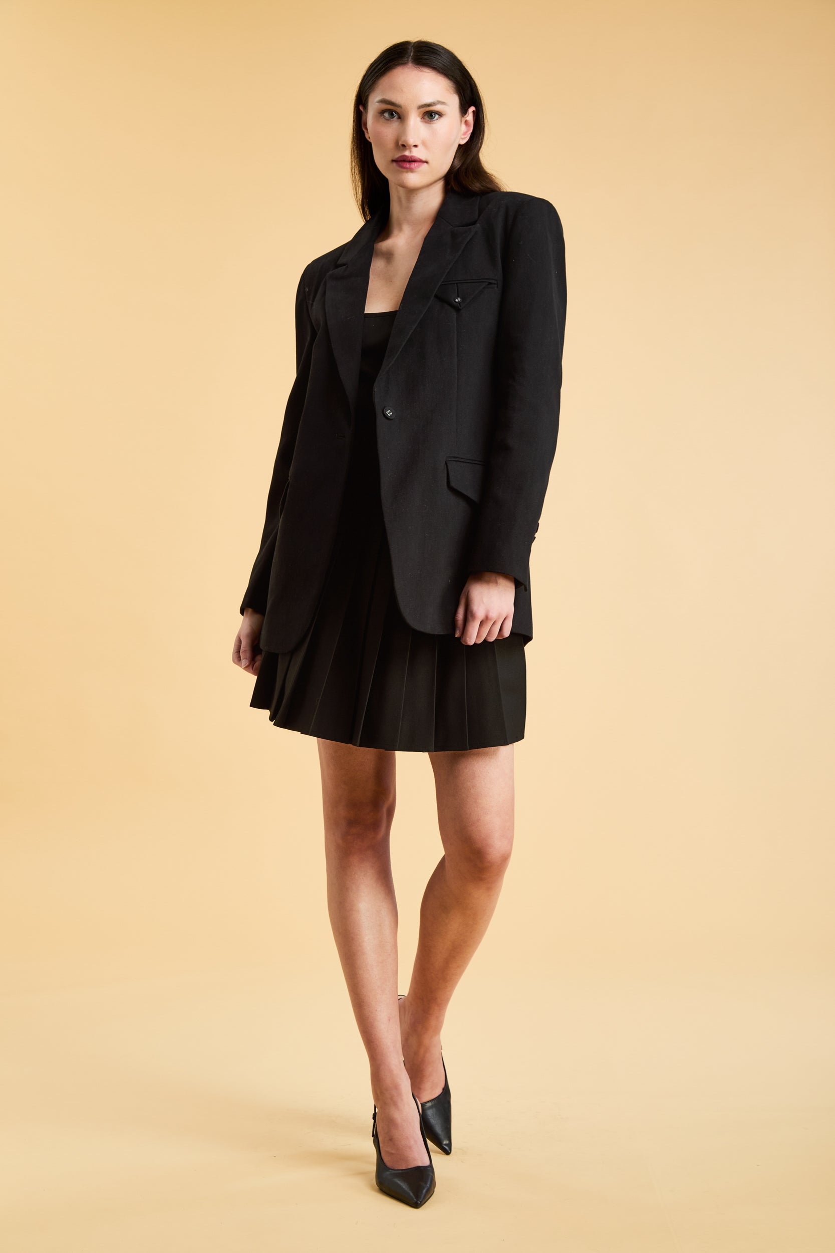Woman wearing a Black oversized tailored Triplicity blazer over a black pleated skirt on a beige background full body