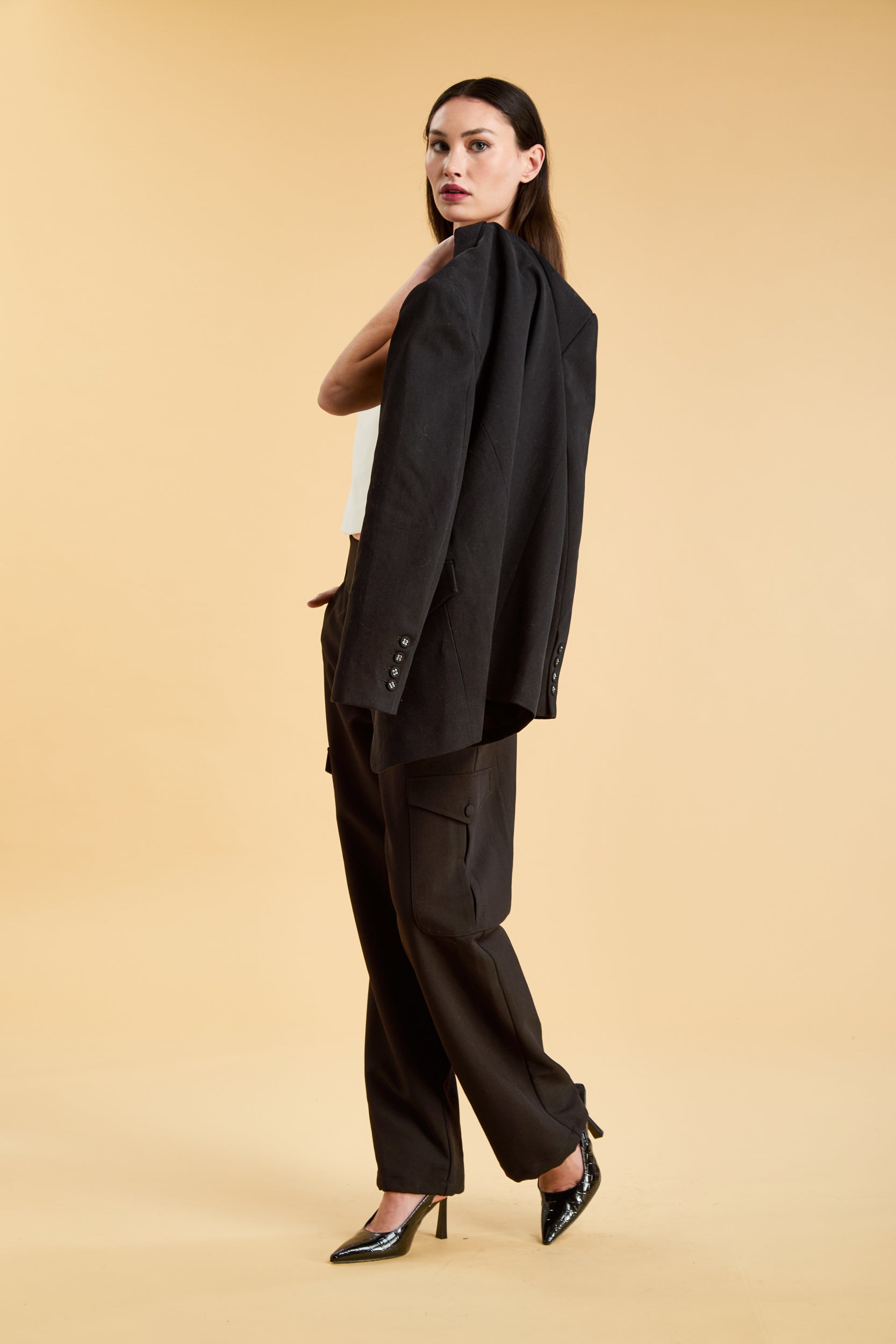 Woman holding a Black oversized triplicity blazer over her shoulder while wearing a white top on a beige background front view full body