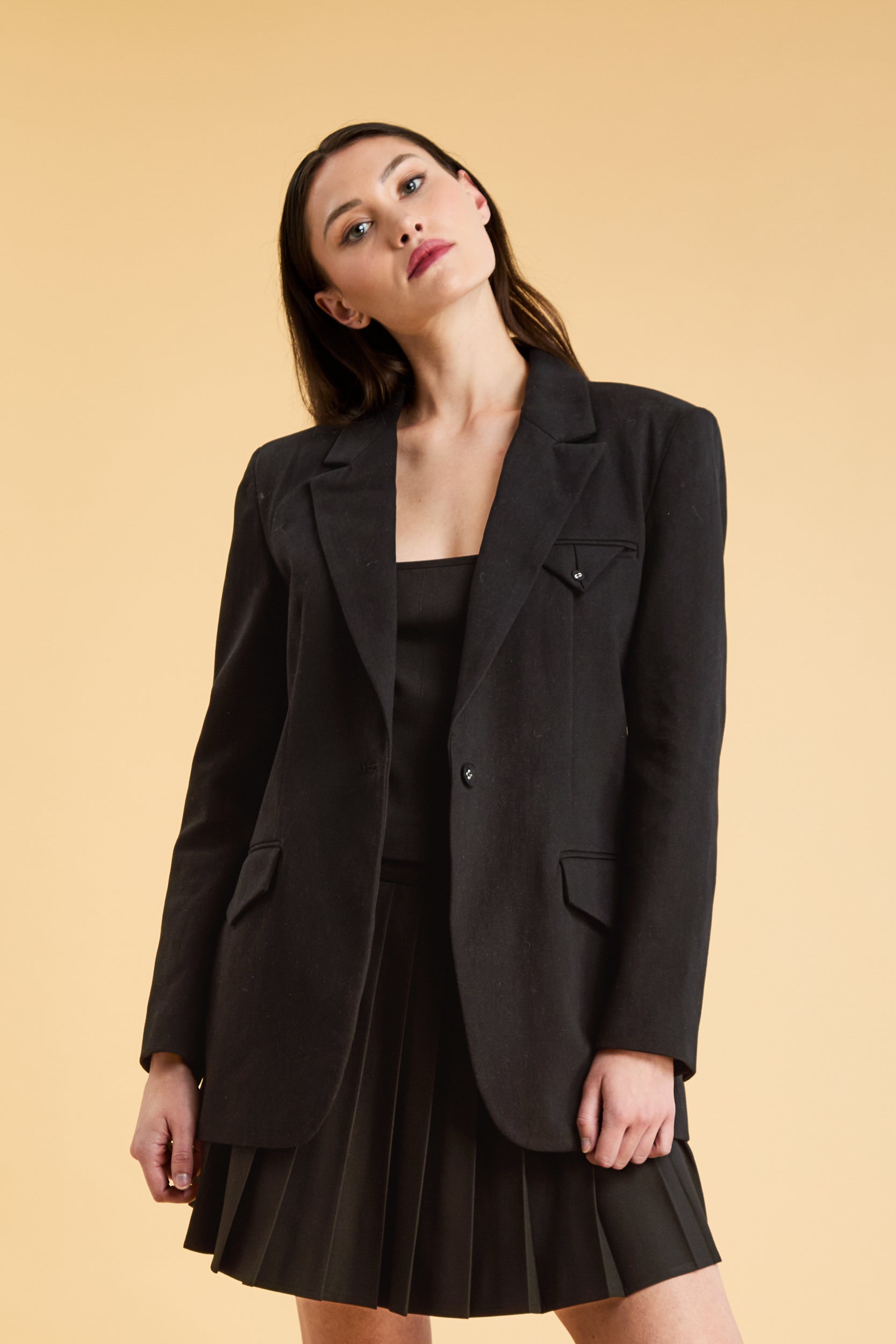 Woman wearing a Black oversized tailored Triplicity blazer over a black pleated skirt on a beige background close up